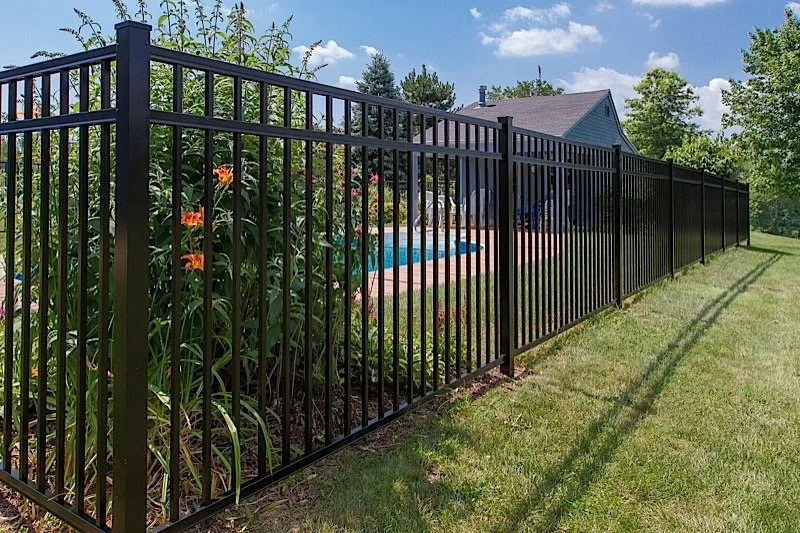 Black metal fence surrounding a backyard with a garden, house, and swimming pool, on a sunny day.