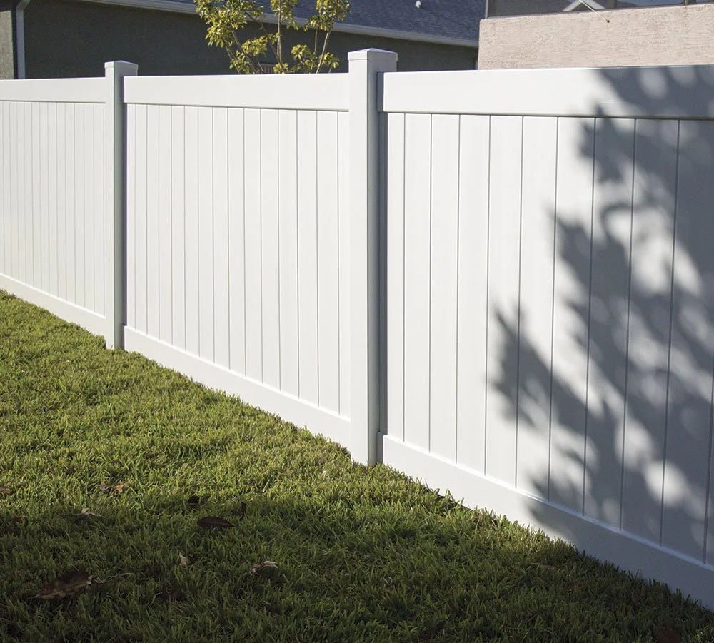 A white privacy vinyl fence with vertical panels and post caps in a residential backyard, casting shadows on the grass.