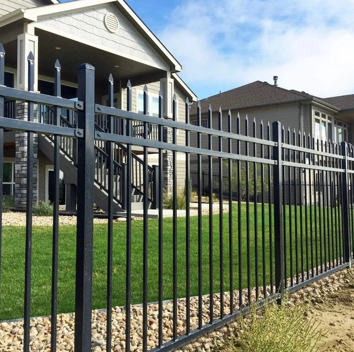 A black metal fence surrounds a green lawn in front of a grey two-story house with a balcony and stairs, under a partly cloudy sky.
