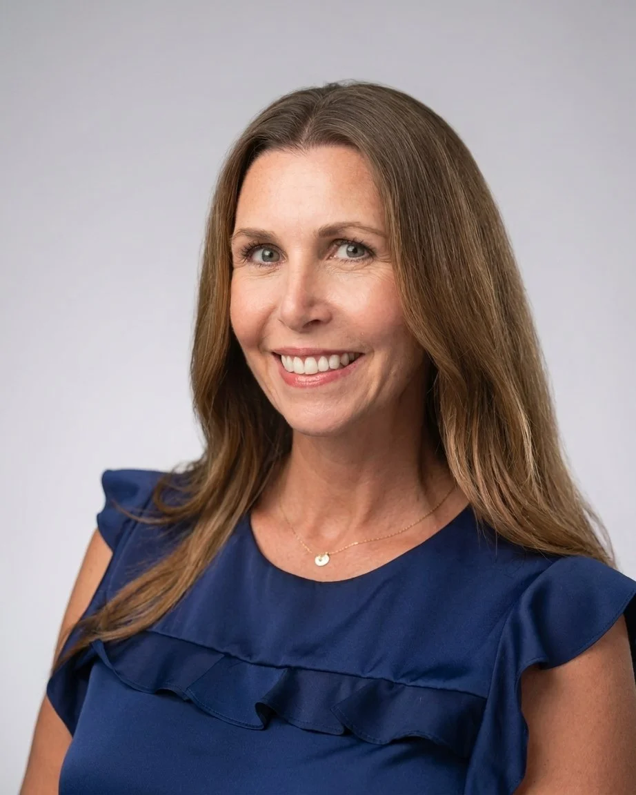 A woman with light skin and long brown hair smiling in a navy blue top with ruffle details, against a plain light gray background.