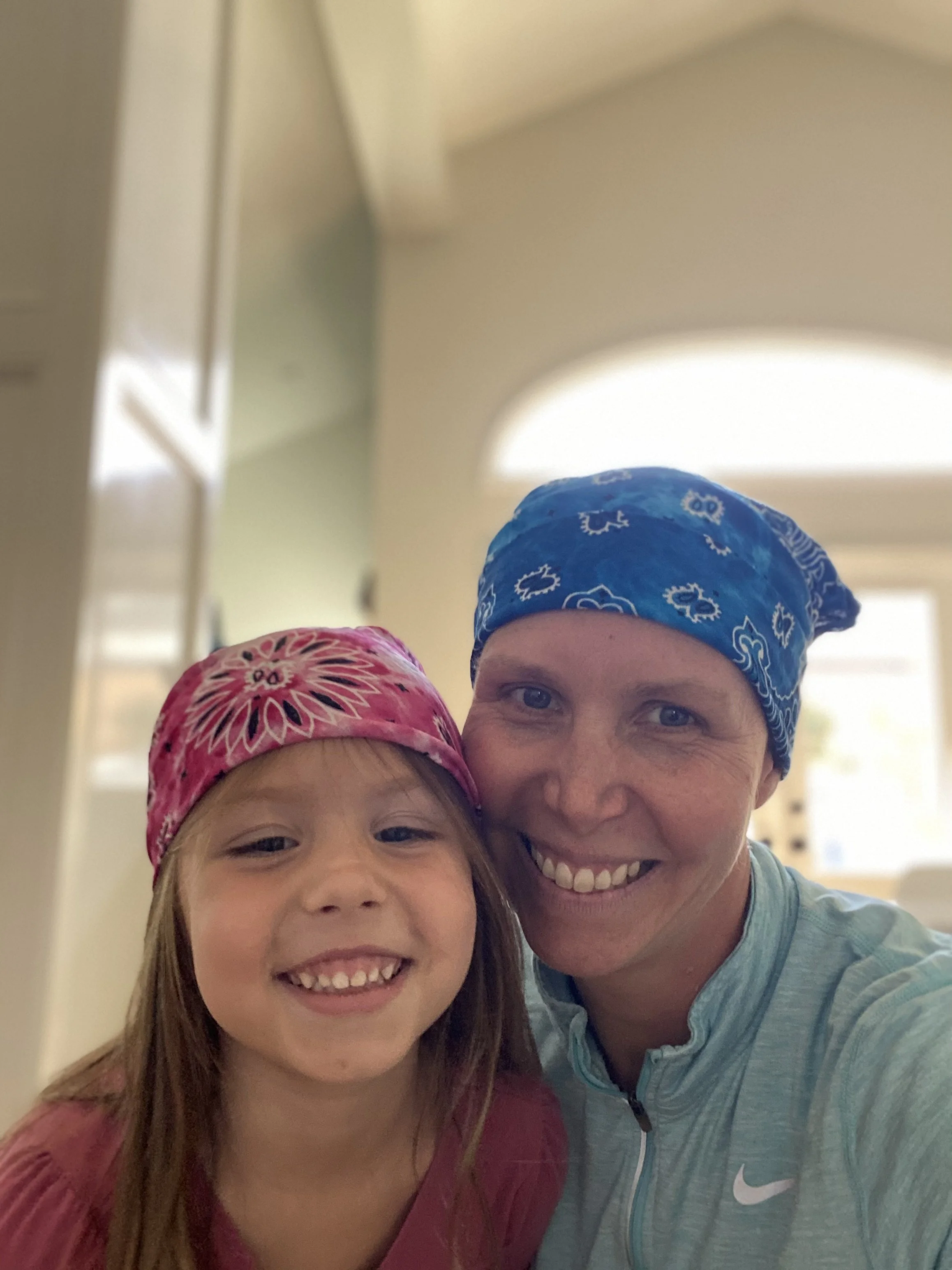 A smiling man and girl taking a close-up selfie indoors, both wearing bandanas on their heads, the girl with a pink bandana and the man with a blue one. The background has a high ceiling with a large window.