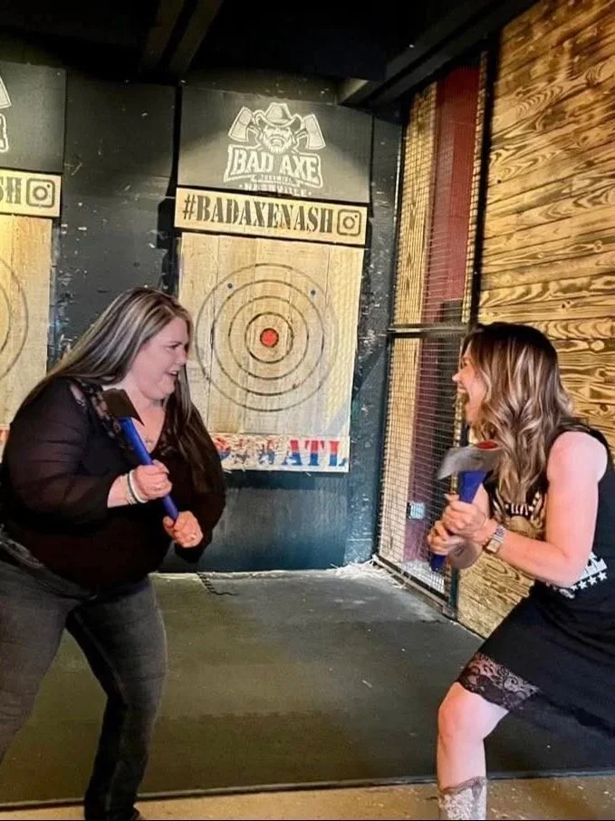 Two women are playing axe throwing at a venue called 'Bad Axe Throwing.' They are smiling and appear to be enjoying the game, each holding axes ready to throw toward a target painted on a wood-paneled wall.