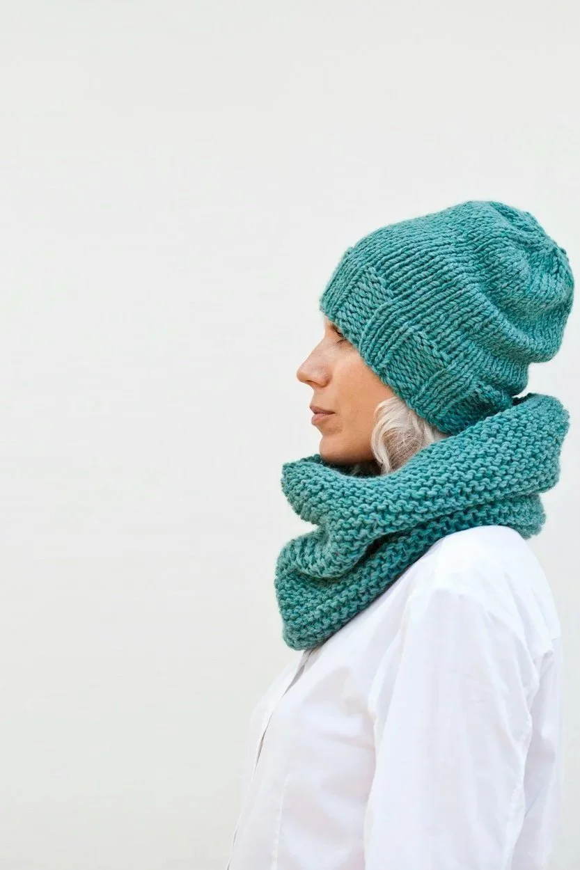 A woman wearing a knit teal beanie and matching knit teal scarf, facing to the left against a plain light background.