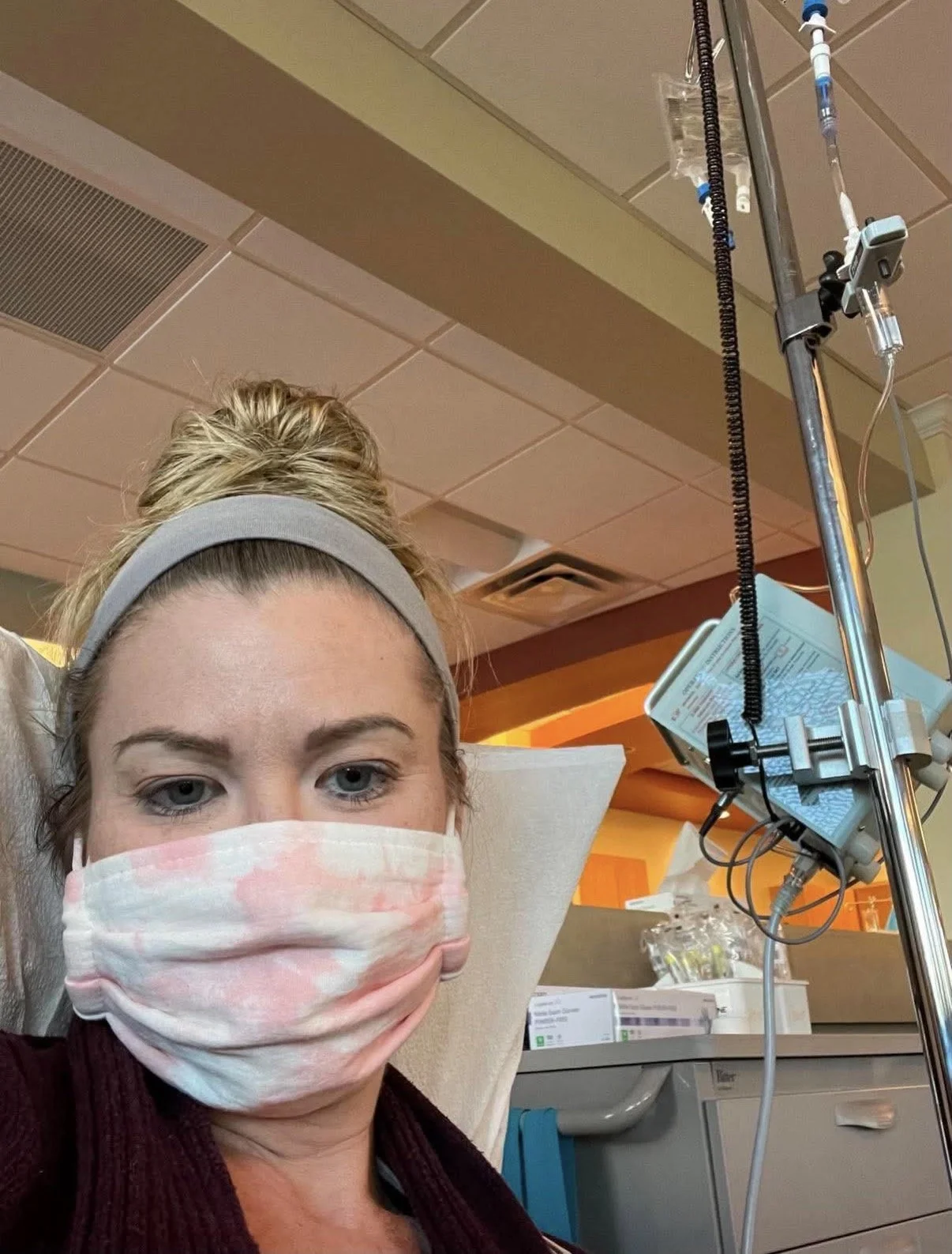 A woman with blonde hair tied in a bun, wearing a gray headband, pink and white face mask, and burgundy sweater, sitting in a hospital room. She is receiving medical treatment with IV equipment and a monitor nearby.