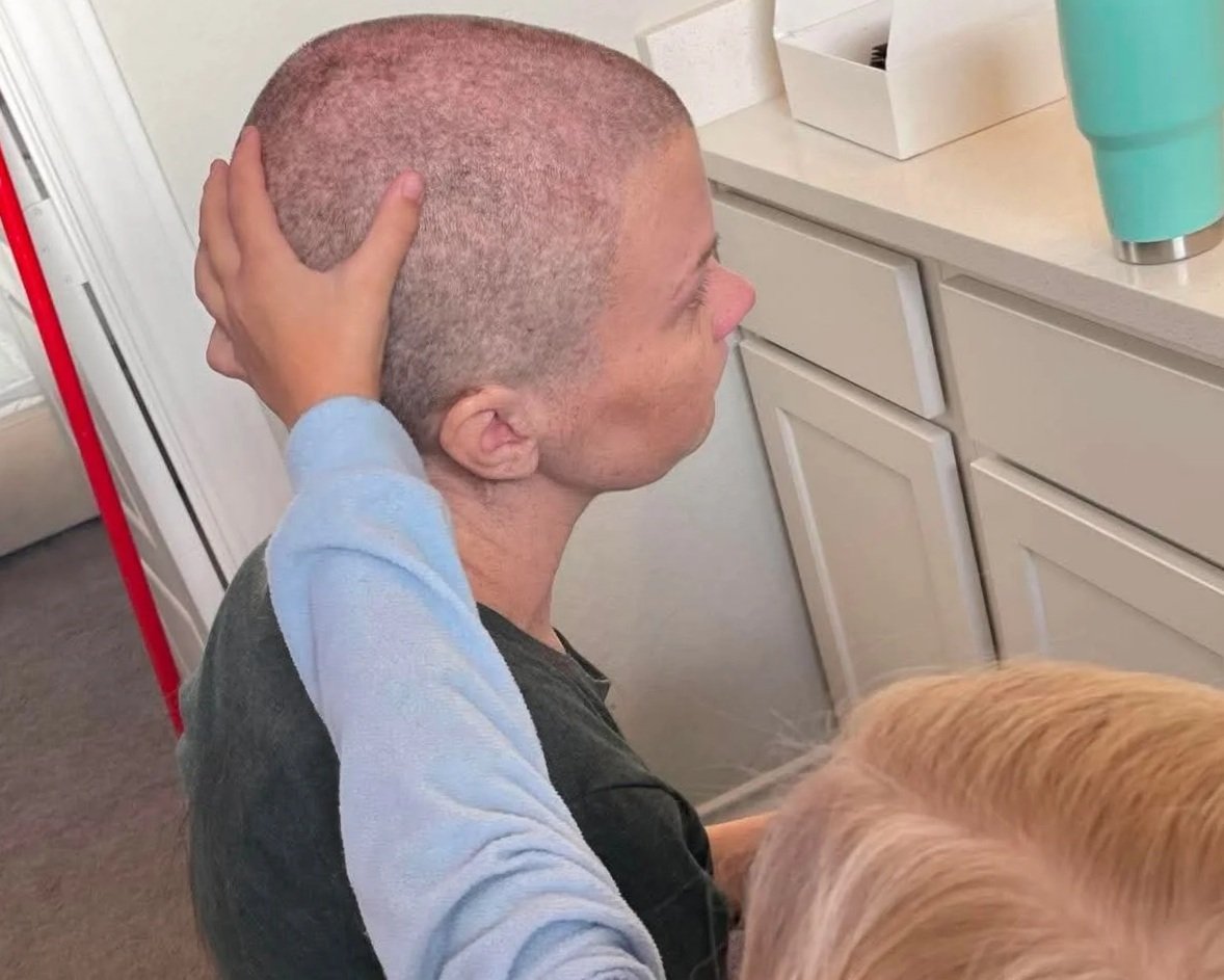 A person with a shaved head is receiving a hand on their head while sitting at a kitchen counter.