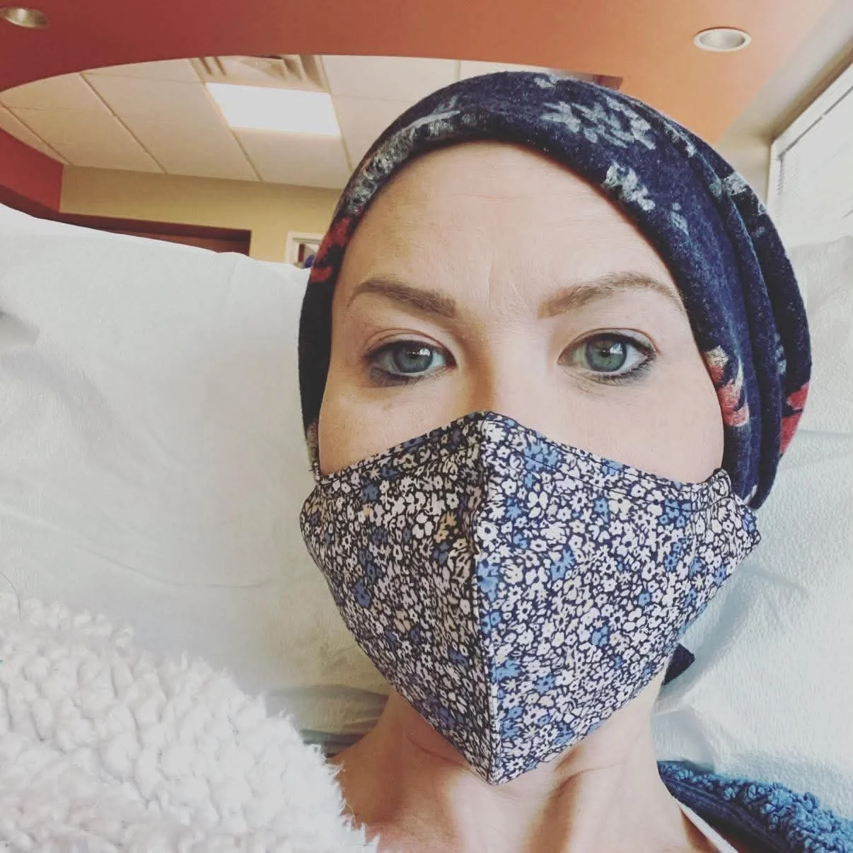 A woman lying in a hospital bed, wearing a patterned fabric face mask and a floral headscarf, with a hospital room background.