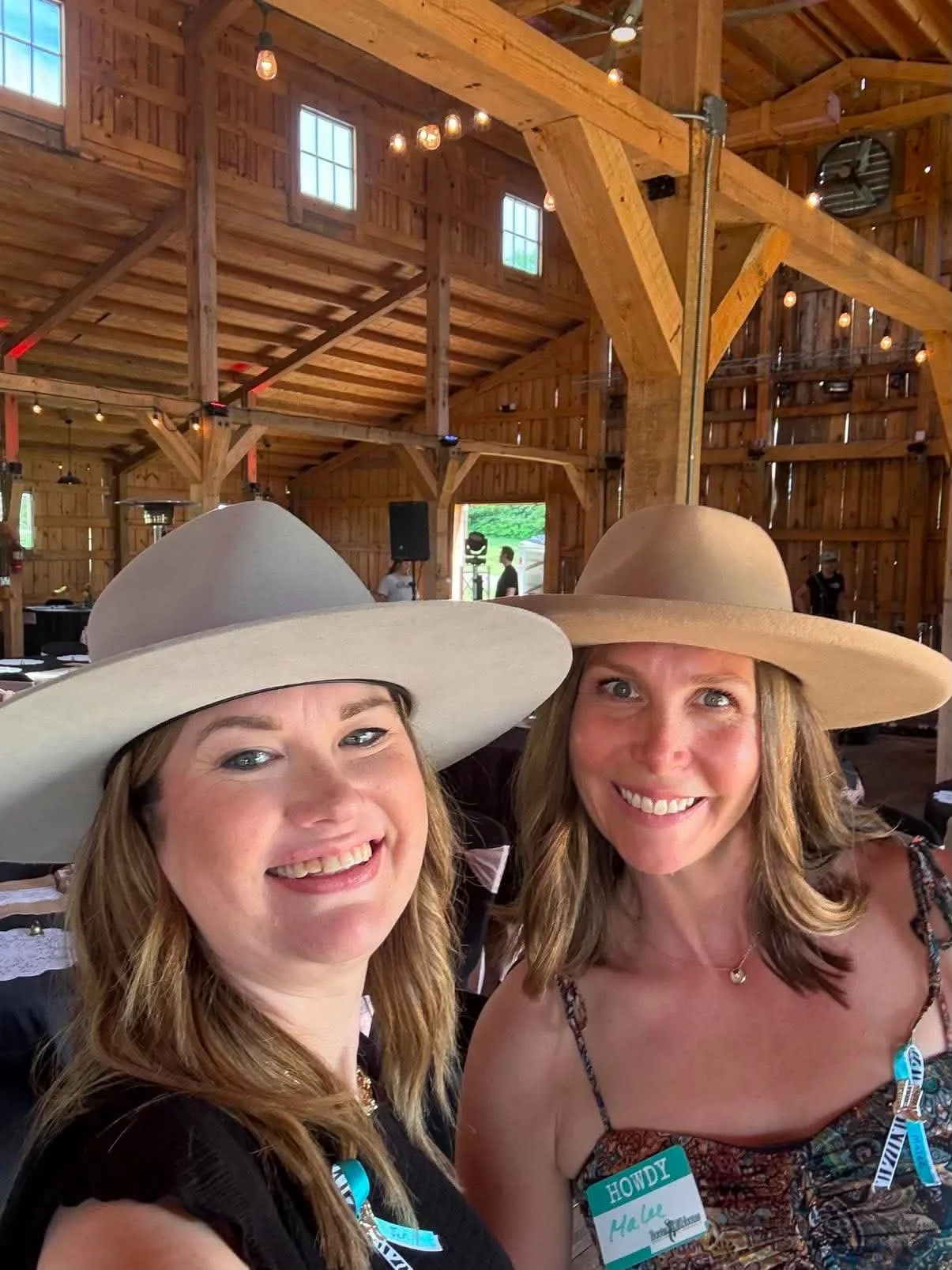 Two women smiling and taking a selfie inside a large wooden barn with high ceilings, wooden beams, and small windows. They are wearing wide-brimmed hats and have name tags, with one reading 'HOWDY'.