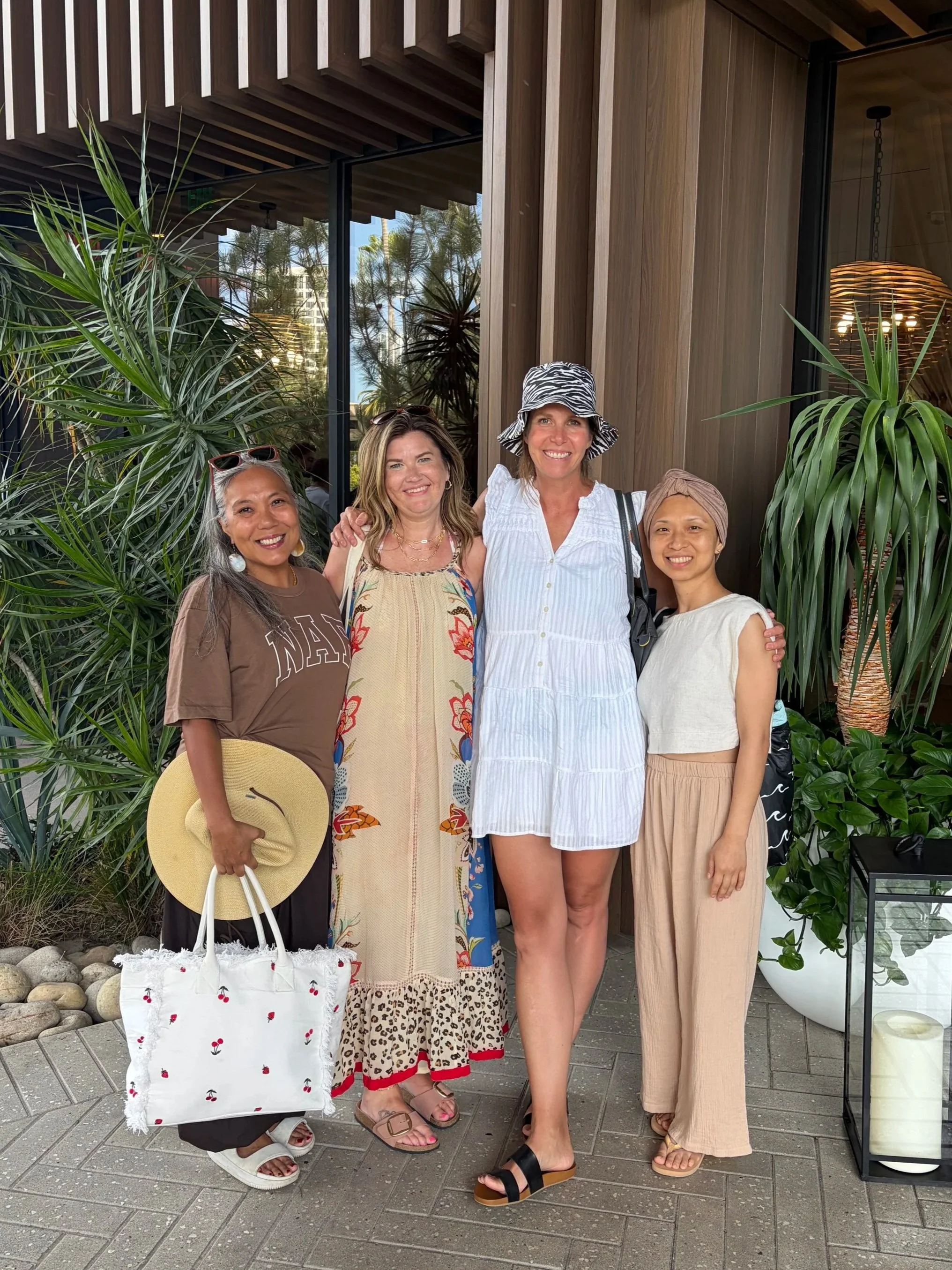 Group of five women standing outdoors near a building with wood paneling and plants. They are smiling and dressed in casual summer clothes. One woman is holding a white tote bag with cherry patterns and a wide-brimmed hat, while another wears a zebra-print sun hat. There are large indoor-style lamps visible inside the building.