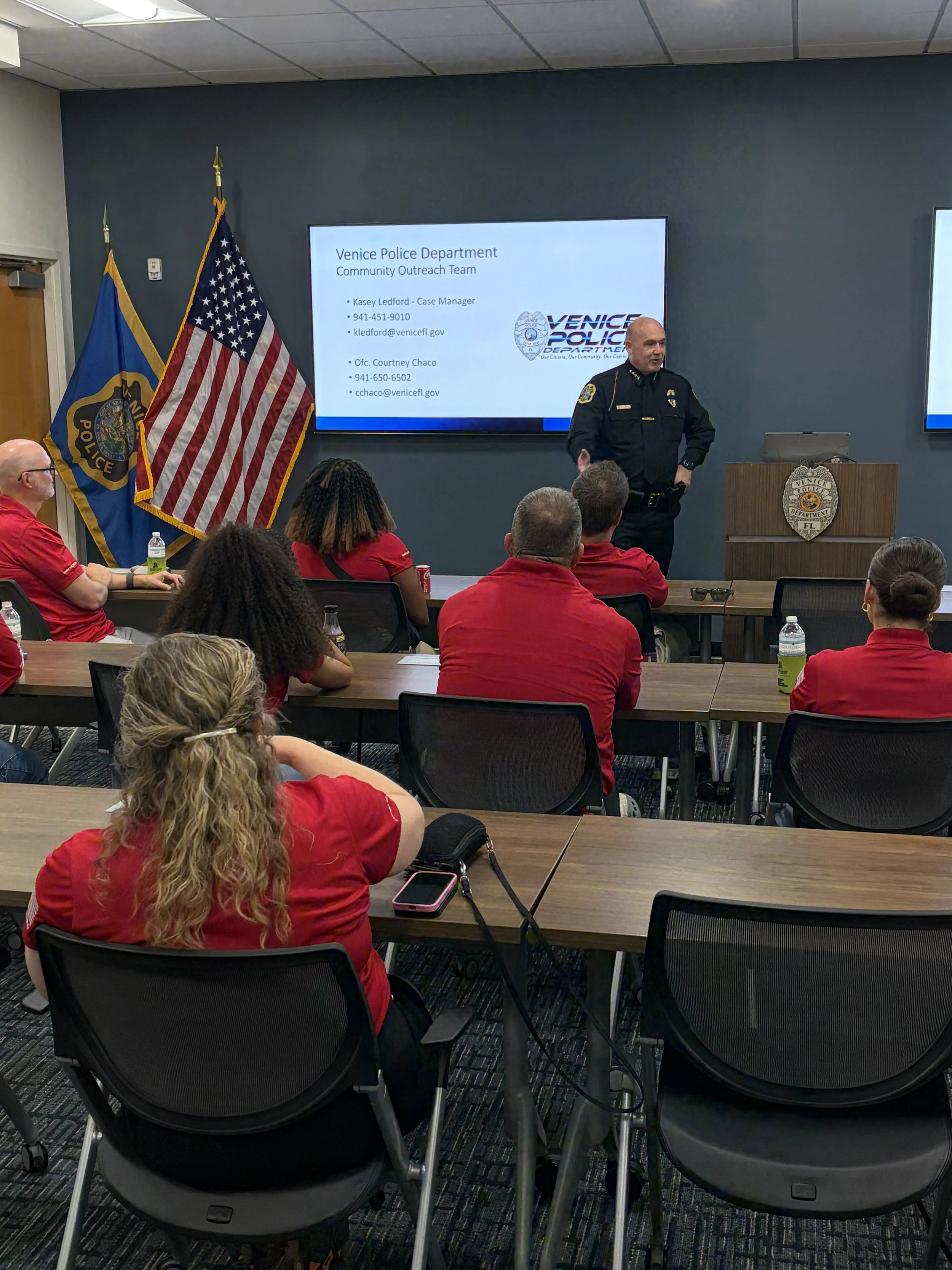 A meeting room with several people, mostly wearing red, attending a presentation by a police officer standing at the front. There are two flags, one American and one of the Venice Police Department, with a mounted monitor displaying a presentation slide titled "Venice Police Department Community Outreach Team."