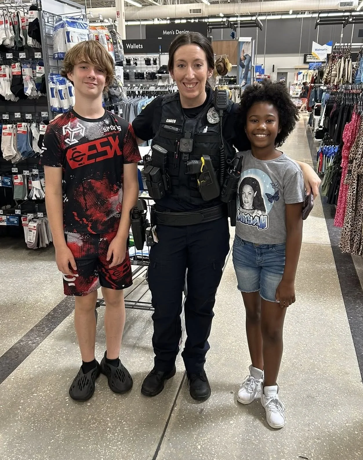 Police officer smiling and standing between two young girls in a store aisle. The girl on the left has short, wavy hair and is wearing a red and black sports outfit. The girl on the right has curly hair, is wearing a gray T-shirt with a graphic and denim shorts. The store has clothing, wallets, and socks visible on shelves and racks.