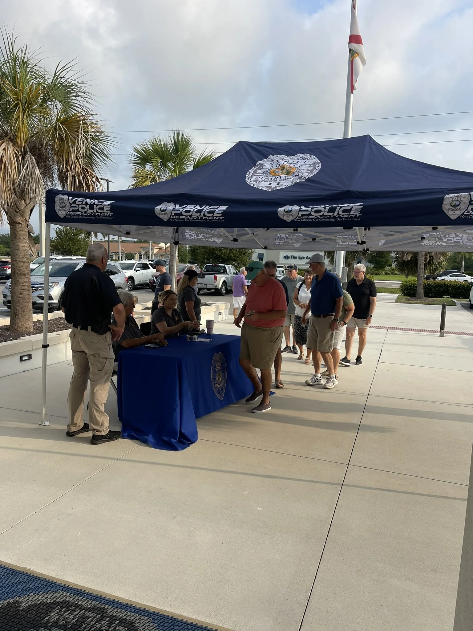 People gathered around a booth under a blue tent with the Venice Police Department logo, outdoors in a parking lot with palm trees, cars, and partly cloudy skies in the background.