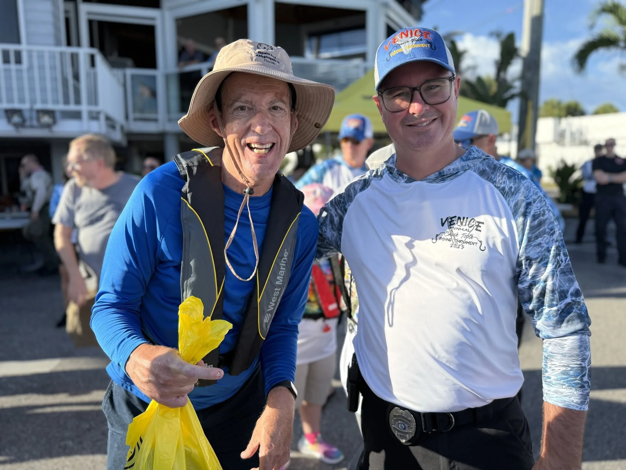 Two men smiling and posing outdoors at a daytime event, with one wearing a wide-brimmed hat and the other wearing a blue cap and glasses. Several people and a building are visible in the background.