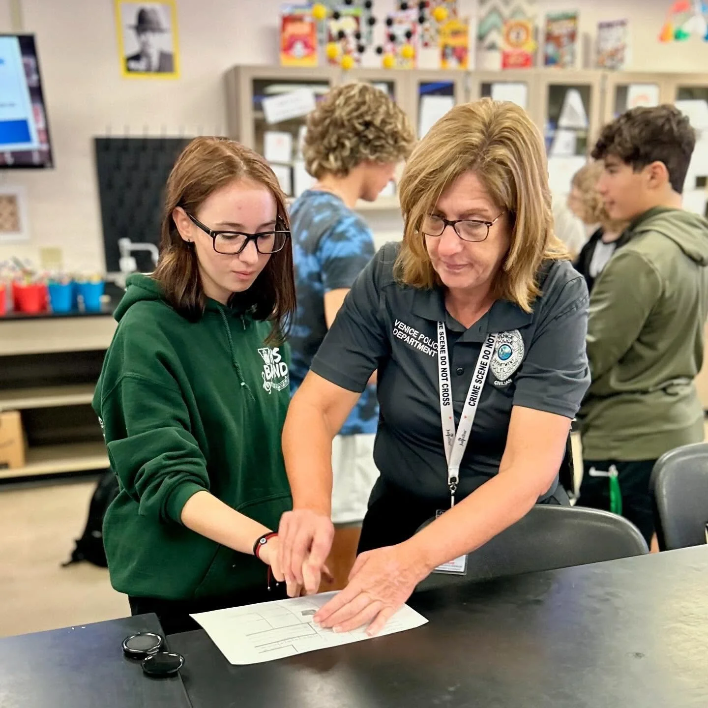 A woman in a police uniform and glasses assists a young girl in a green hoodie with a craft project at a table in a classroom or community center. Several other people are working or walking in the background.