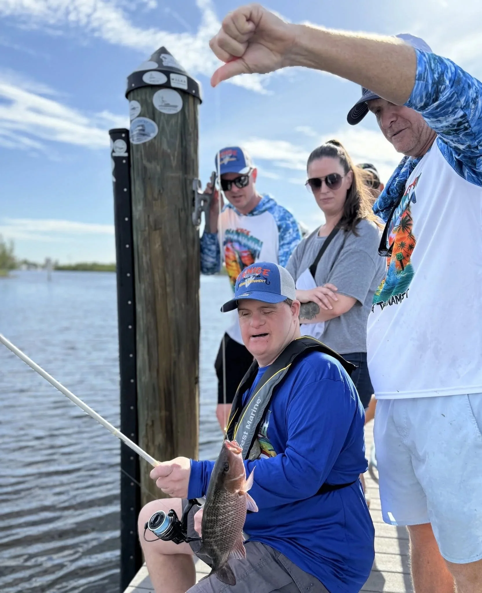 A group of people fishing by a body of water on a sunny day, with a man holding a fish he caught.