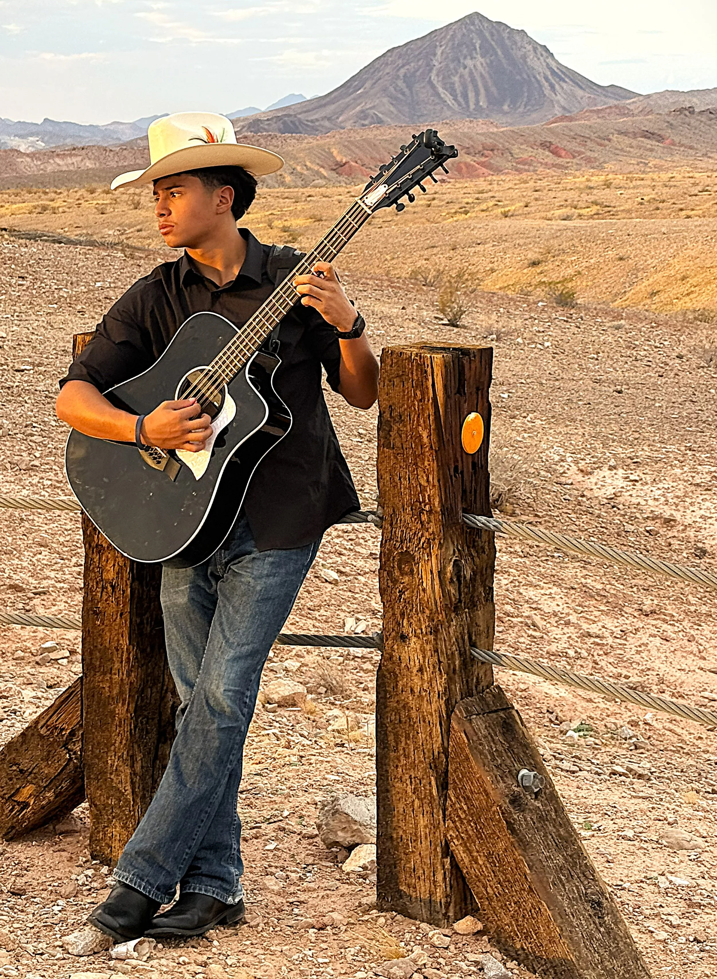 Young man wearing a cowboy hat playing an acoustic guitar outdoors, with a mountain in the background. Rancho, Ranch.