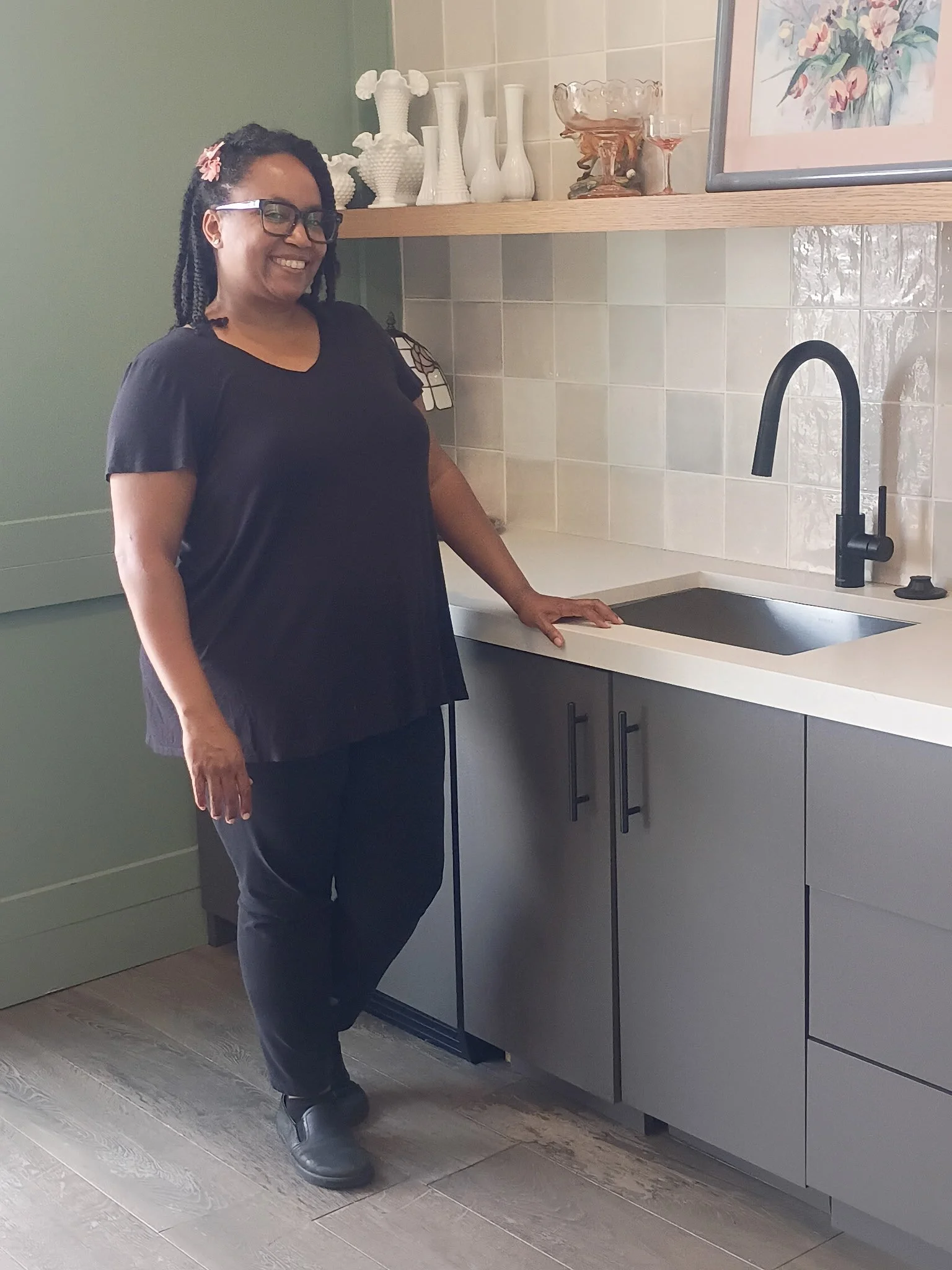 A woman with glasses and dreadlocks smiling in a modern kitchen, standing next to a black sink and blue-gray cabinets, with decorative vases and a framed floral painting on the wall.