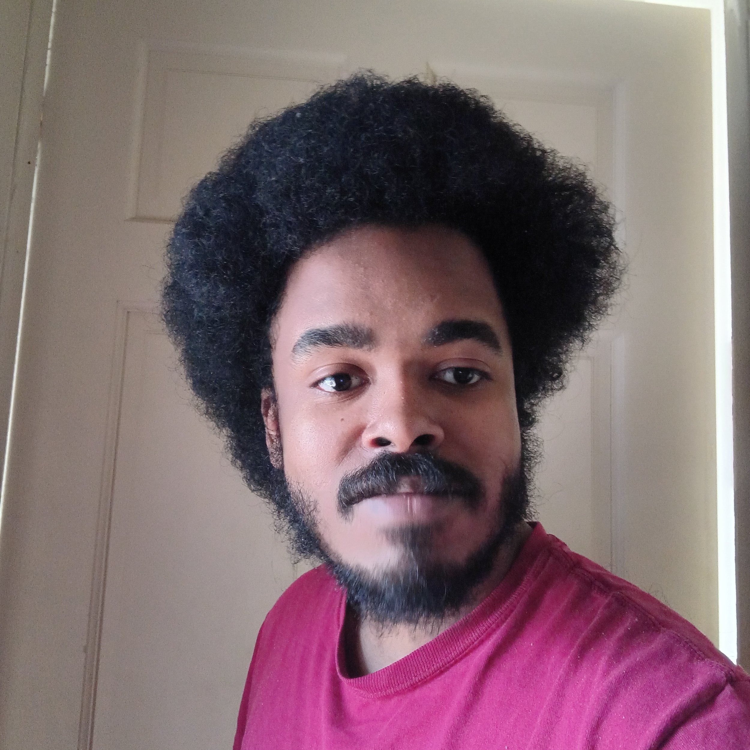 Close-up of a person's face with dark curly hair, facial hair, and wearing a red shirt.
