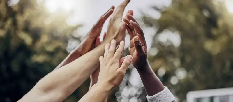 Multiple hands reaching upwards in a high-five or group gesture outdoors with trees in the background.