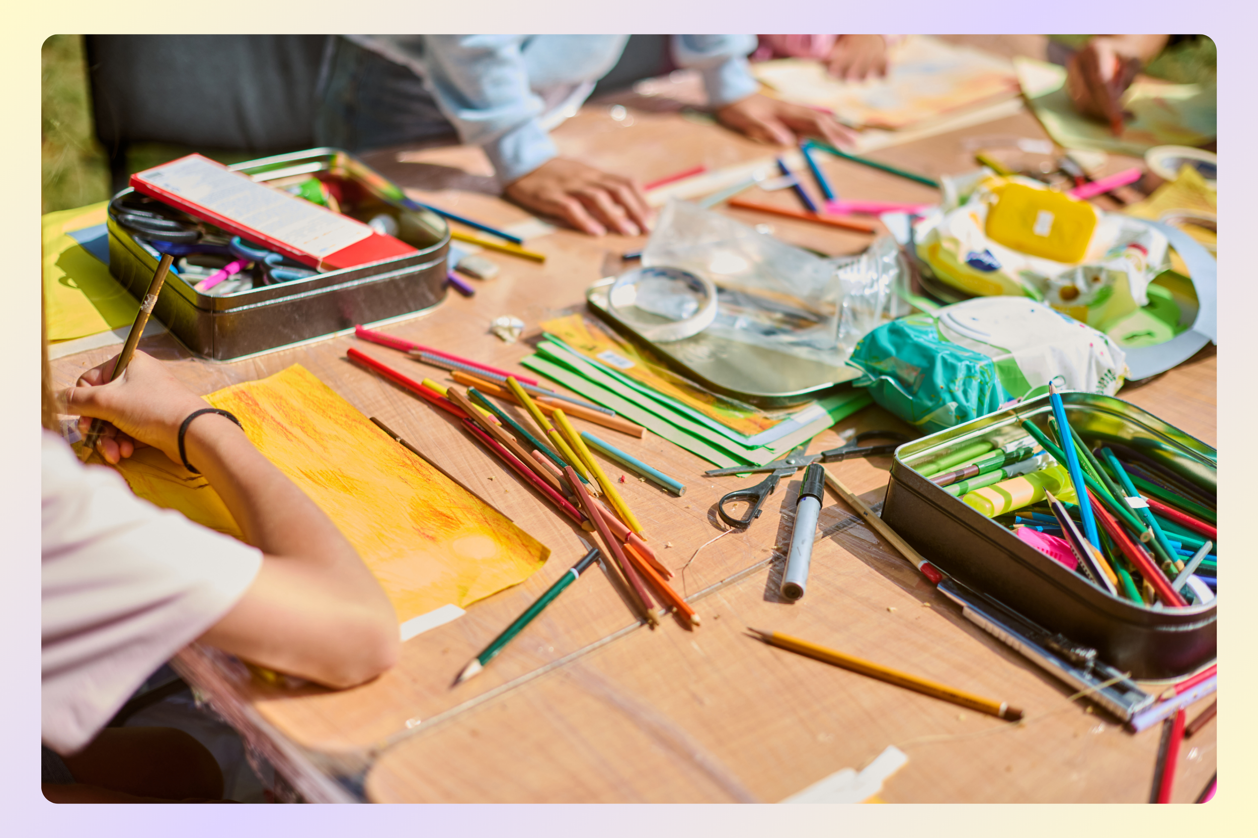 Children's hands drawing with colored pencils on yellow paper at a table filled with art supplies.
