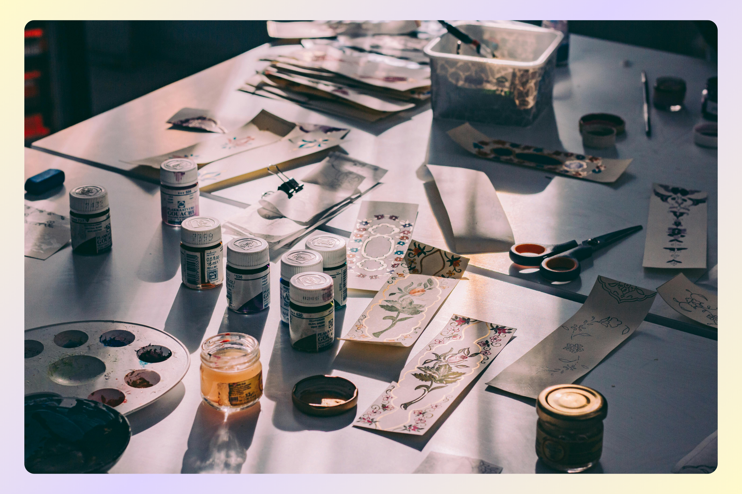 An art workspace featuring watercolor paints, brushes, paper sheets with floral and decorative designs, scissors, and containers, illuminated by natural light.