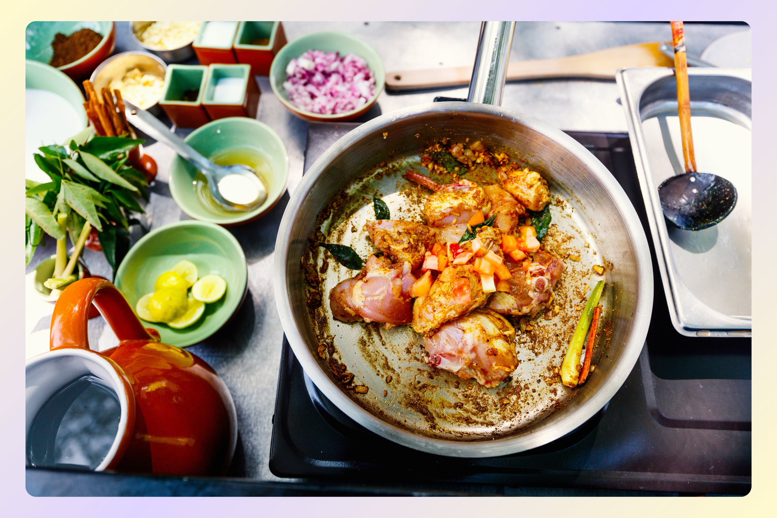 A cooking scene showing chicken pieces in a pan with spices and vegetables, surrounded by various bowls of ingredients and vegetables on a kitchen counter.