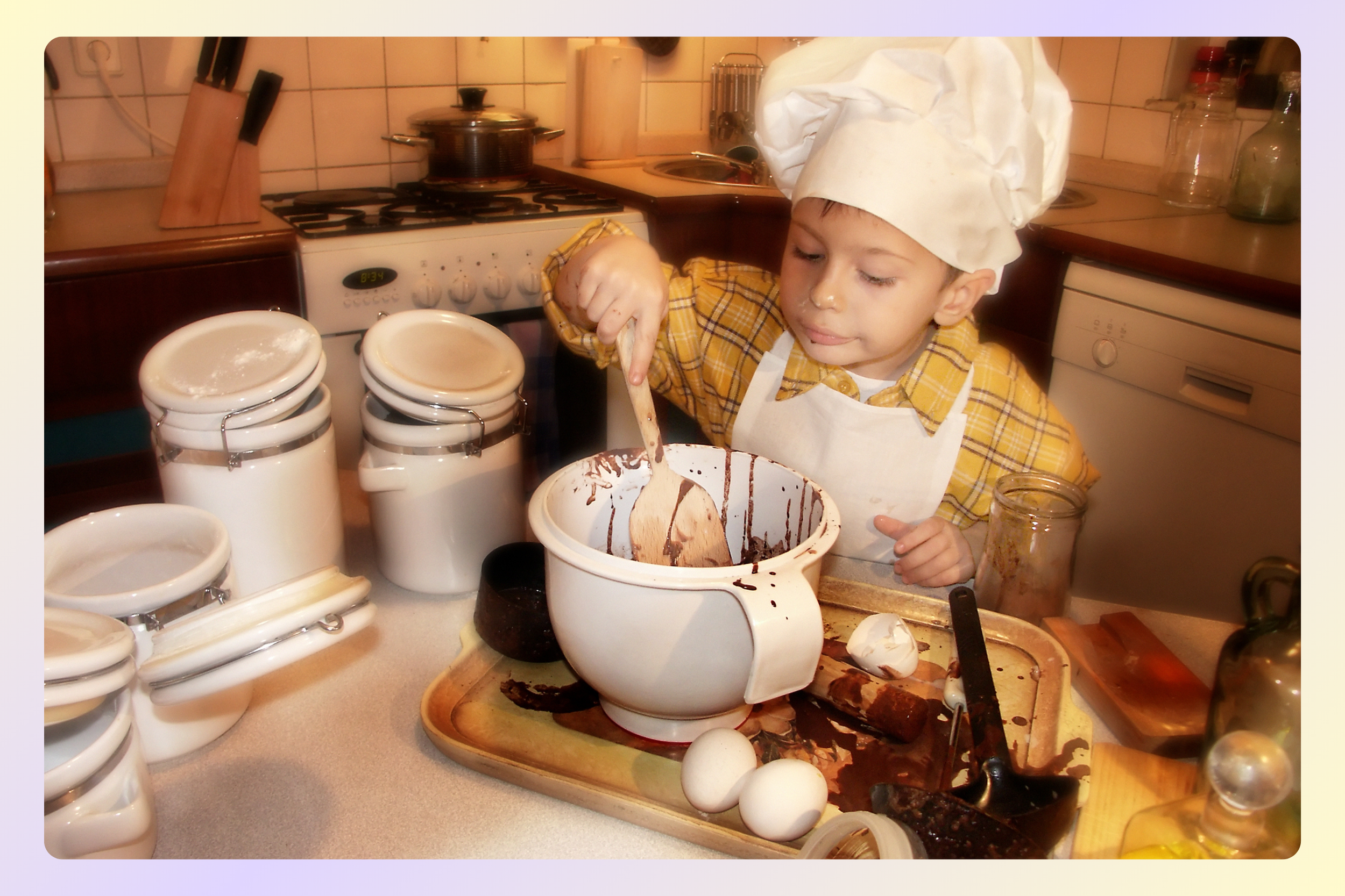 A young child wearing a chef's hat and apron preparing a chocolate dessert in a kitchen. The child is stirring a mixture in a white pot with chocolate drips visible. The countertop is scattered with eggs, jars, and kitchen utensils, and there are stacks of empty ceramic containers nearby.