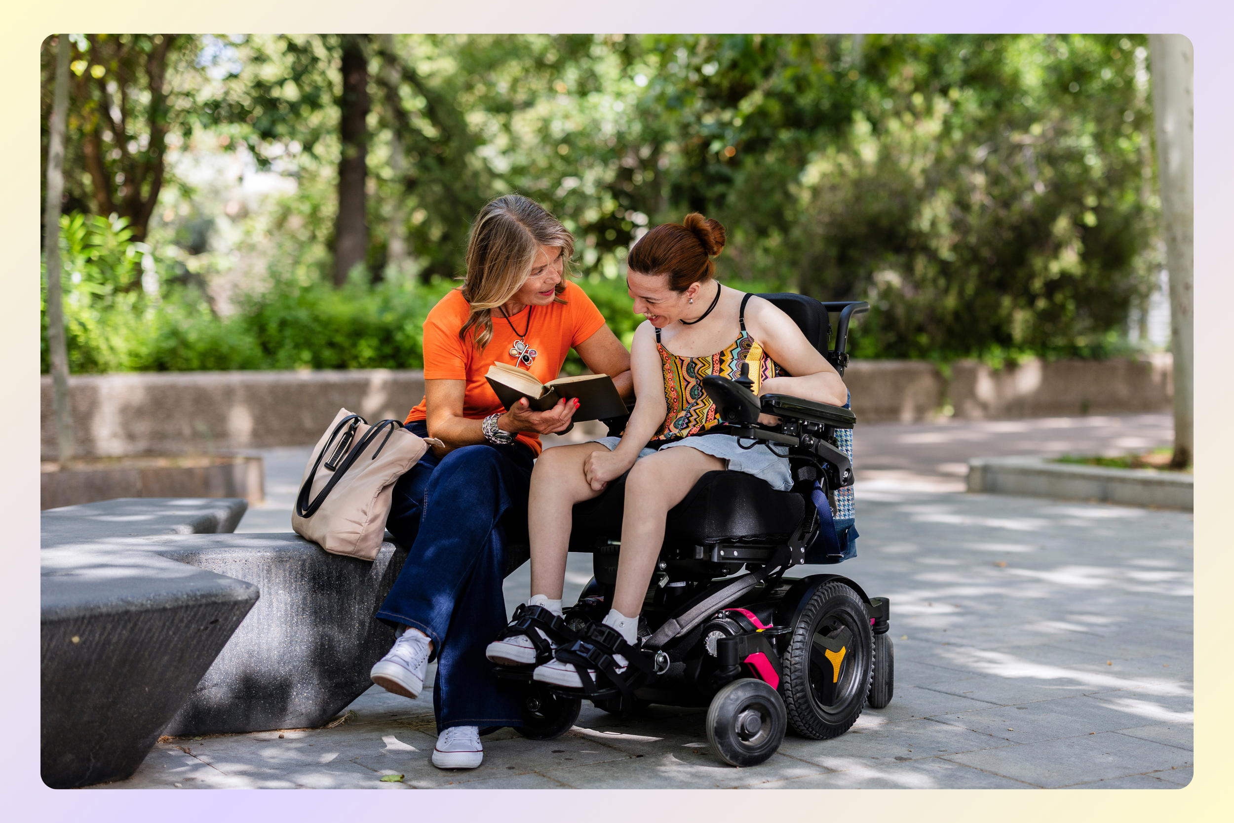 A woman in an orange shirt and a woman in a floral top in a wheelchair sitting on a park bench, sharing a book and smiling at each other with a background of green trees.
