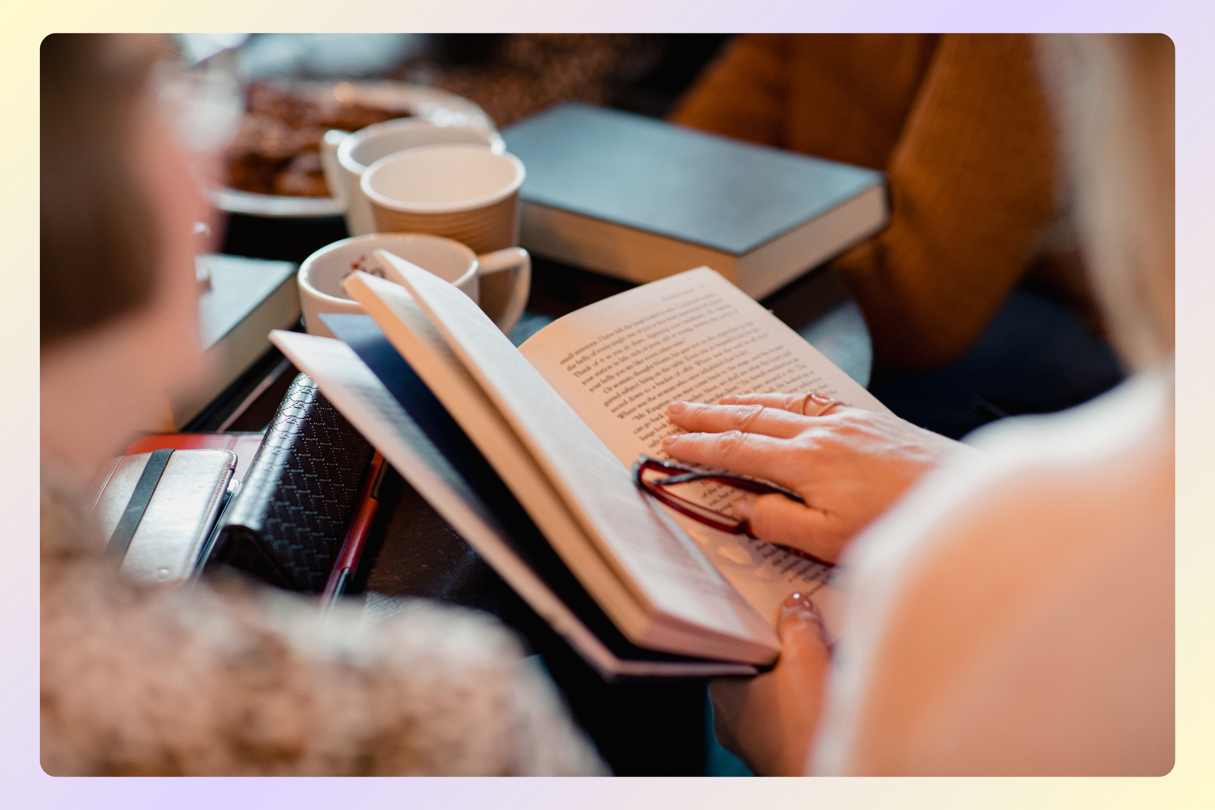 A person reading a book at a table with coffee mugs, notebooks, and pens.
