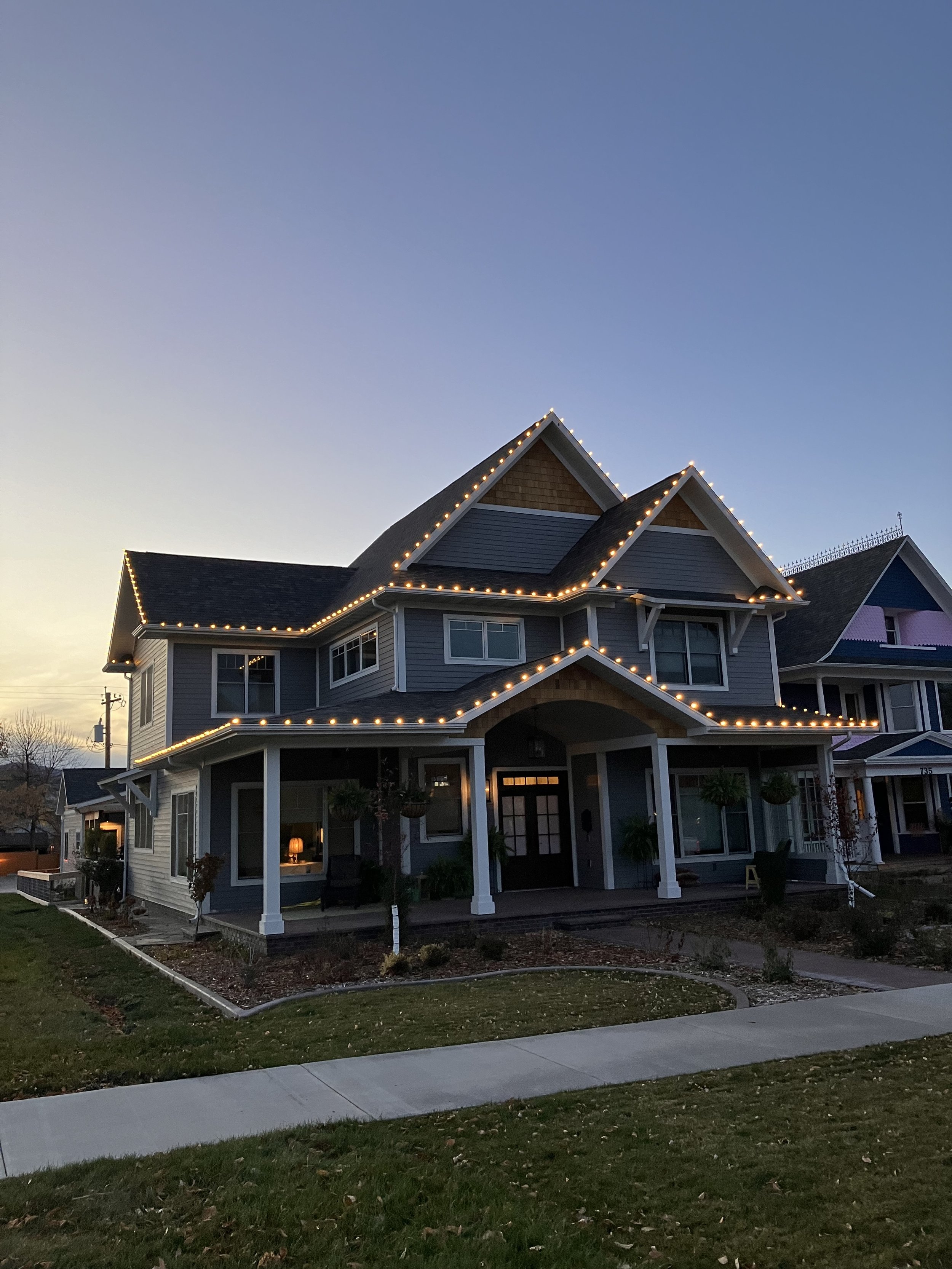 Two story home at dusk with warm white Christmas lights outlining the roofline and porch