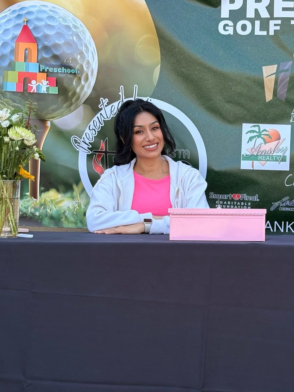 A smiling woman with long black hair, wearing a pink shirt and a white jacket, sitting at a table with her arms crossed. Behind her is a promotional backdrop with logos and images, including a golf ball, a preschool graphics, a palm tree, and boxing gloves, with some flowers in a vase on her left.