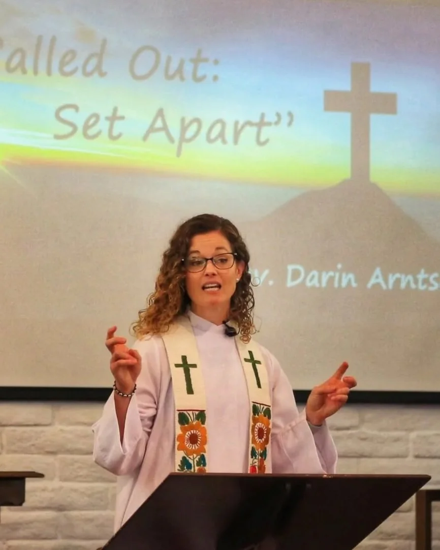 Rev. Darin Arntson, with glasses and brown curly hair, wearing a white robe and a multi-colored cross stole, giving a sermon at her church.