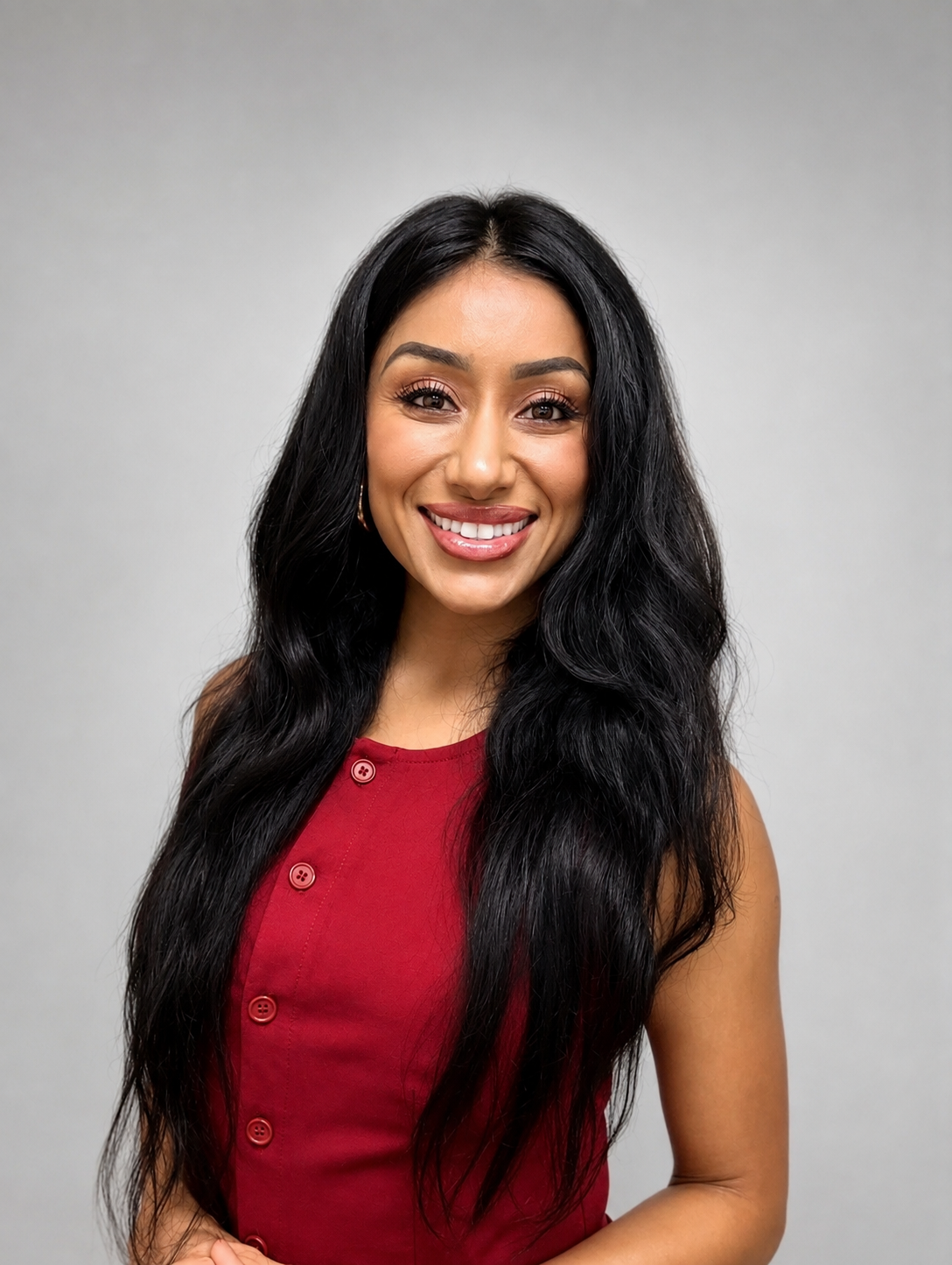Portrait of a smiling woman with long black hair wearing a red sleeveless top with buttons, standing against a plain light gray background.