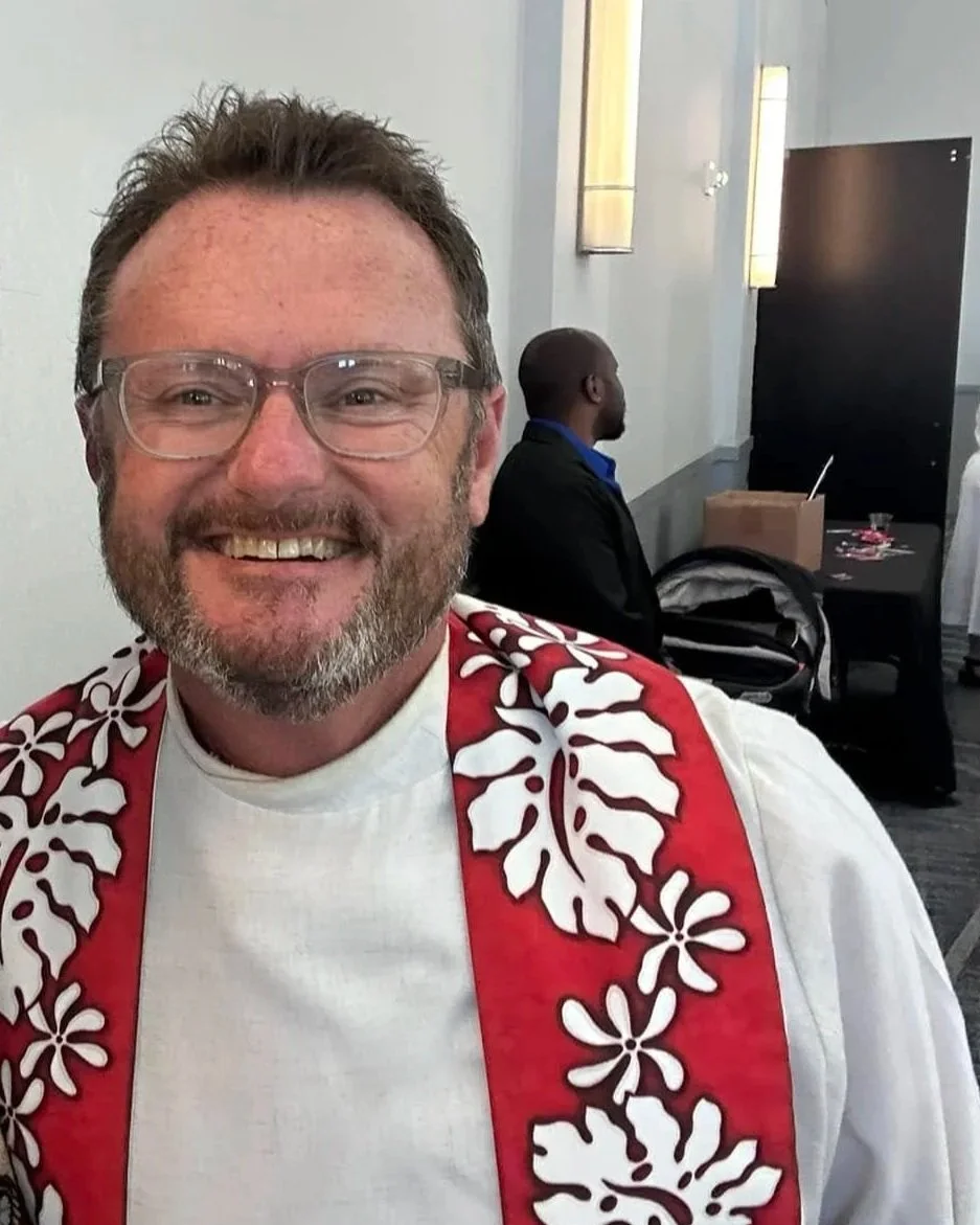 A smiling man, Rev. Brian Timothy Parcel, with glasses and a beard, wearing a white shirt with a red and white floral-patterned sash, sitting at a conference or event with another man in the background.