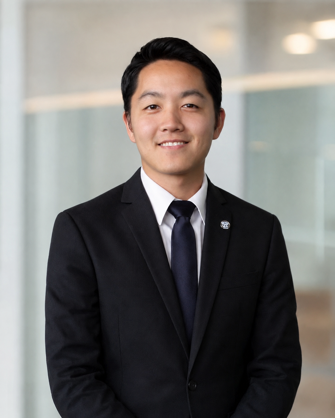 A young man in a black suit, white shirt, and navy tie, smiling in a professional setting with blurred background.