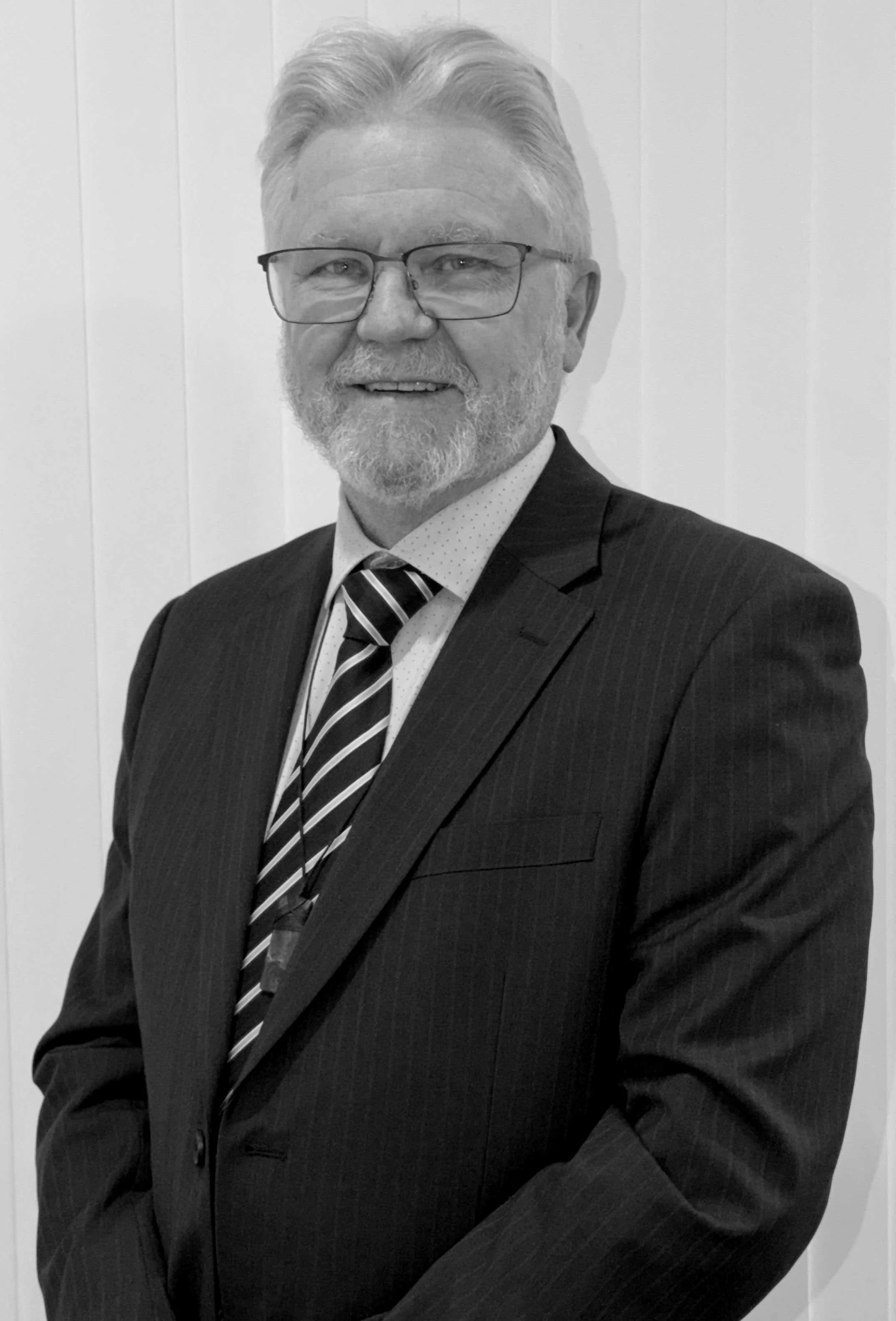 A black and white photo of a middle-aged man with white hair, glasses, and a beard, dressed in a dark pinstripe suit, white shirt, and striped tie, smiling and standing in front of a plain wall.