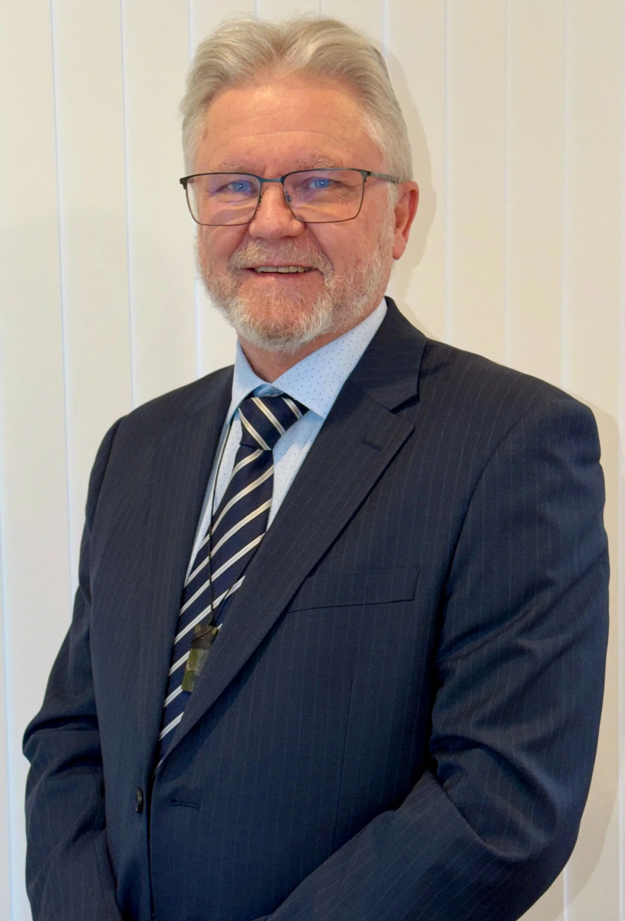 Headshot of an older man with gray hair, beard, wearing glasses and a dark pinstripe suit, white dress shirt, and striped tie, standing against a cream-colored background.