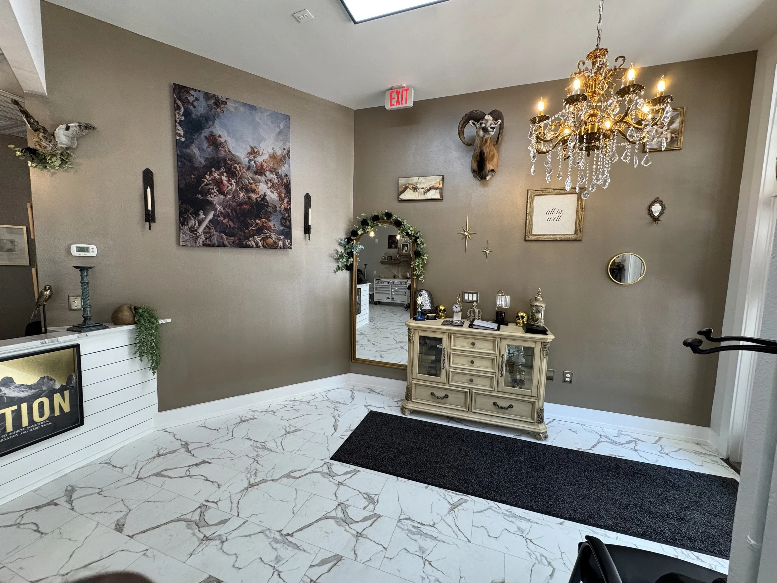 A foyer with marble flooring, a chandelier, a wall-mounted ram's head, framed artwork, and decorative items on a cream-colored side table.