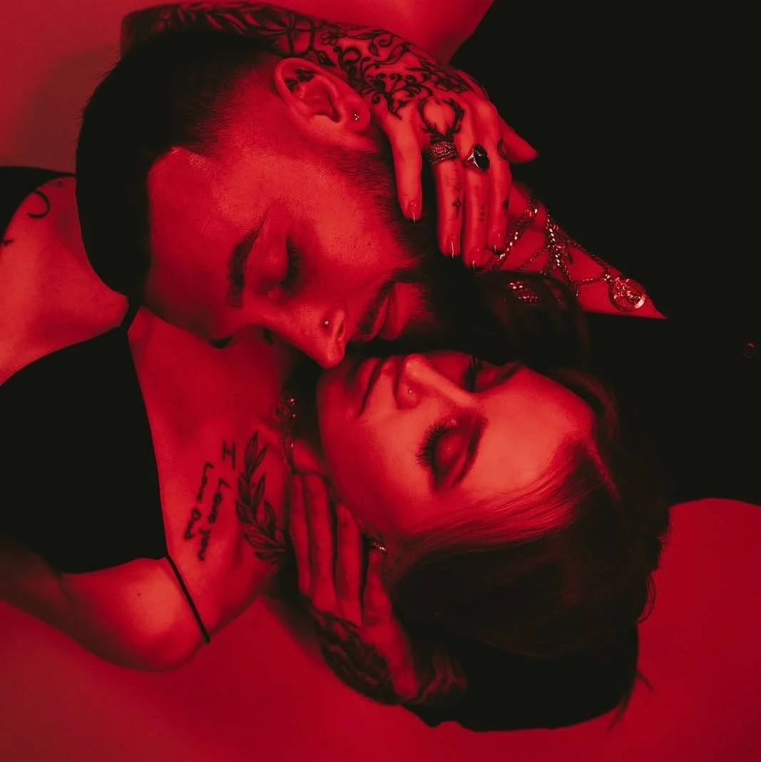 A man with tattoos and jewelry and a woman with tattoos and earrings, lying closely together in a red-hued scene, with their eyes closed and faces touching.