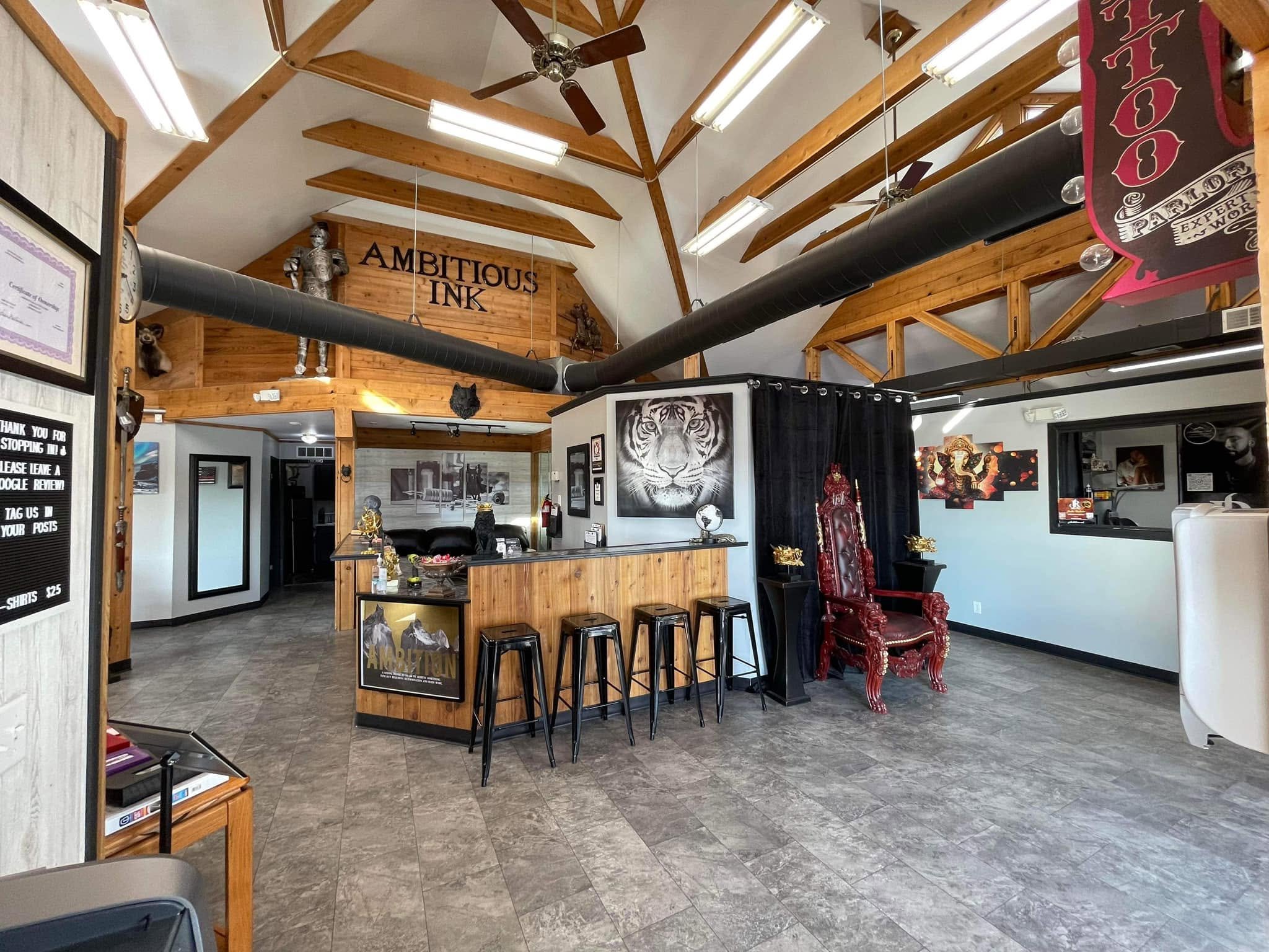Interior of a tattoo shop with wooden beams and a sign that says "Ambitious Ink" on the back wall. There is a tattoo sign on a black curtain, black stools at a bar counter, and artwork including a tiger on the wall. A decorative red throne-style chair and various artwork and signage are also visible.