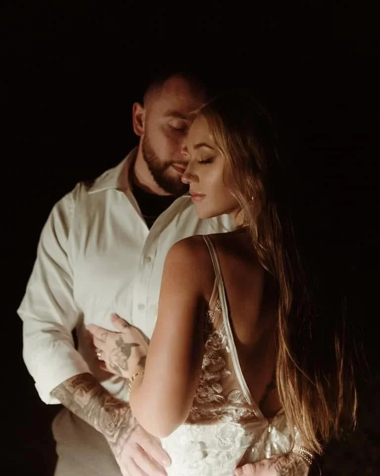A man and woman in an intimate pose, with their foreheads touching. The woman has long hair and is wearing a satin, lace dress. The man has a beard, tattoos on his arms, and is wearing a white shirt. They are set against a dark background.