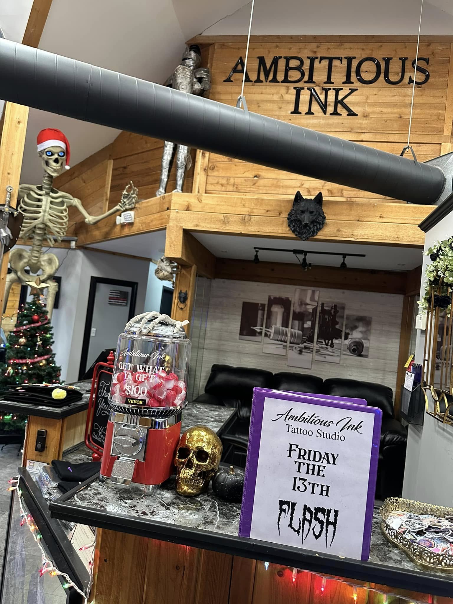 Decorations and signage at a tattoo studio, including a skeleton wearing a Santa hat, a black wolf head sculpture, Halloween-themed skulls, a small Christmas tree with ornaments, a transparent container with pink and red candy, and an event sign for Friday the 13th. The background features a wooden wall with the words 'Ambitious Ink'.