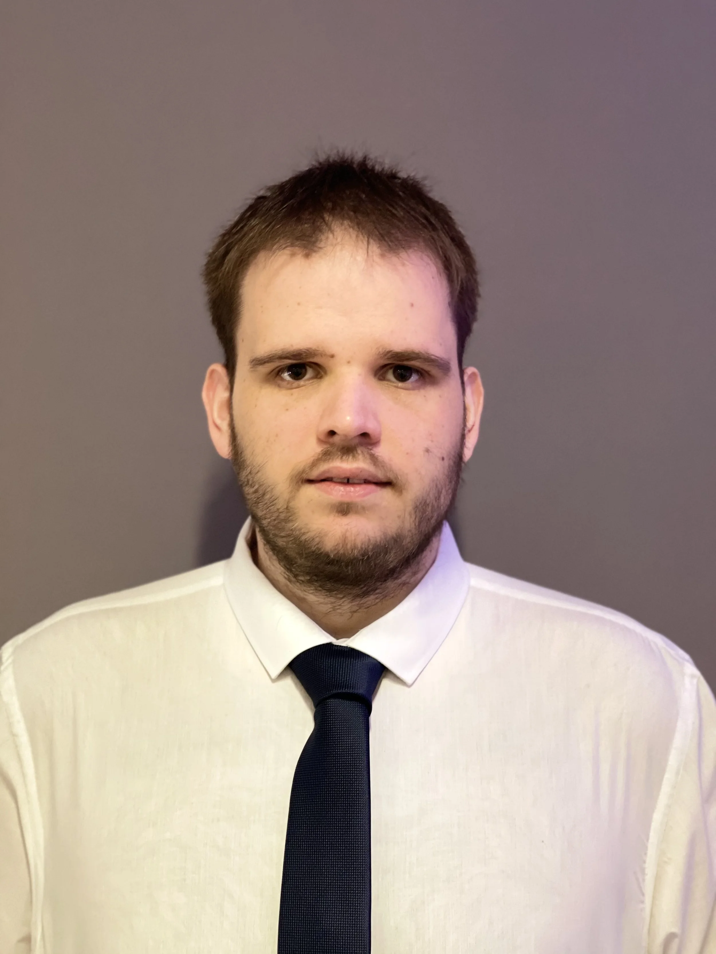 Young man with short brown hair and a beard, wearing a white dress shirt and dark tie, standing against a plain gray background.