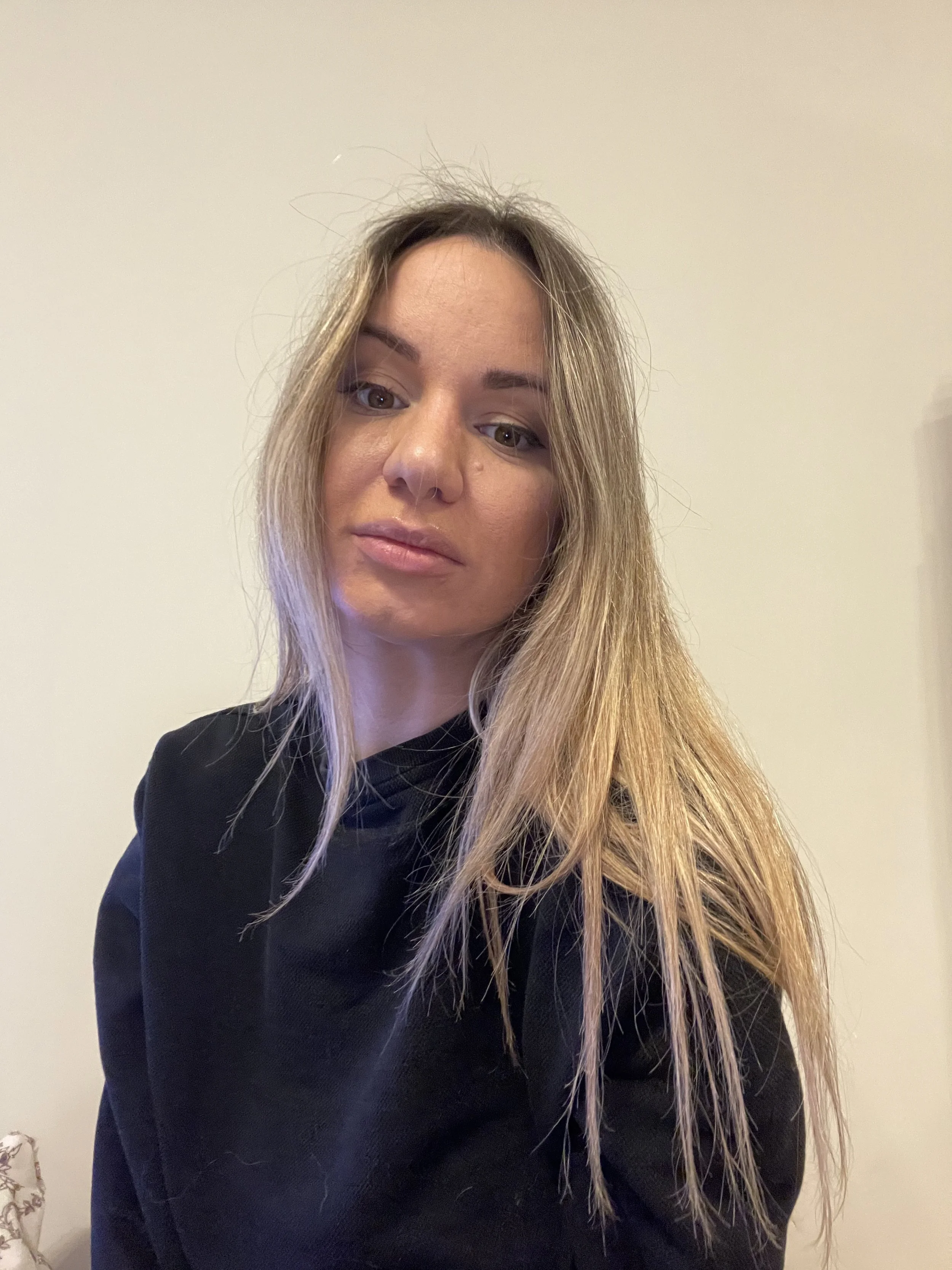 A woman with long blonde hair and light makeup, wearing a black shirt, standing against a plain light-colored wall.
