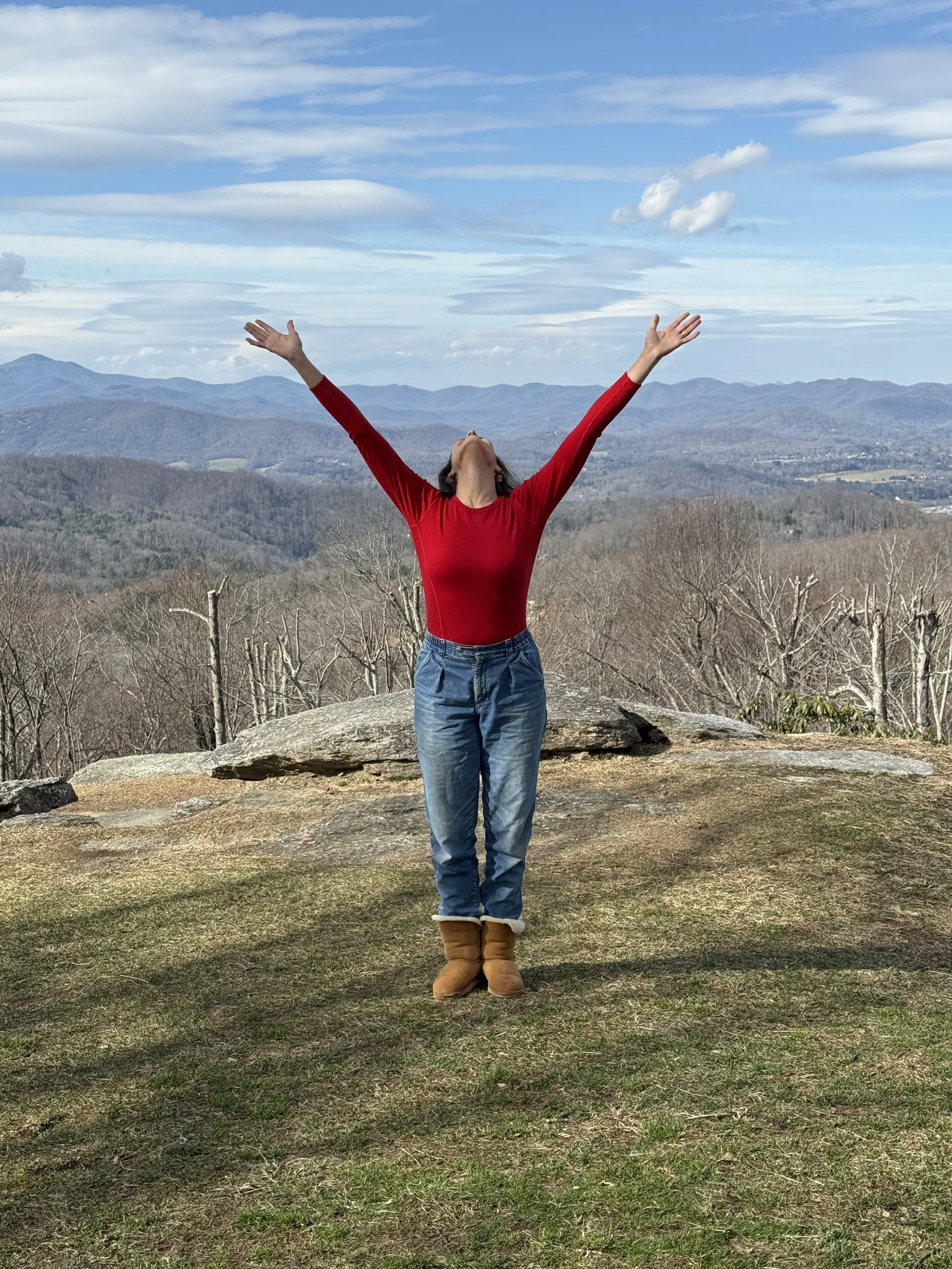 A person standing outdoors on a hilltop with arms raised, facing away, wearing a red long-sleeve top, blue jeans, and brown boots, with a mountain landscape and cloudy sky in the background.