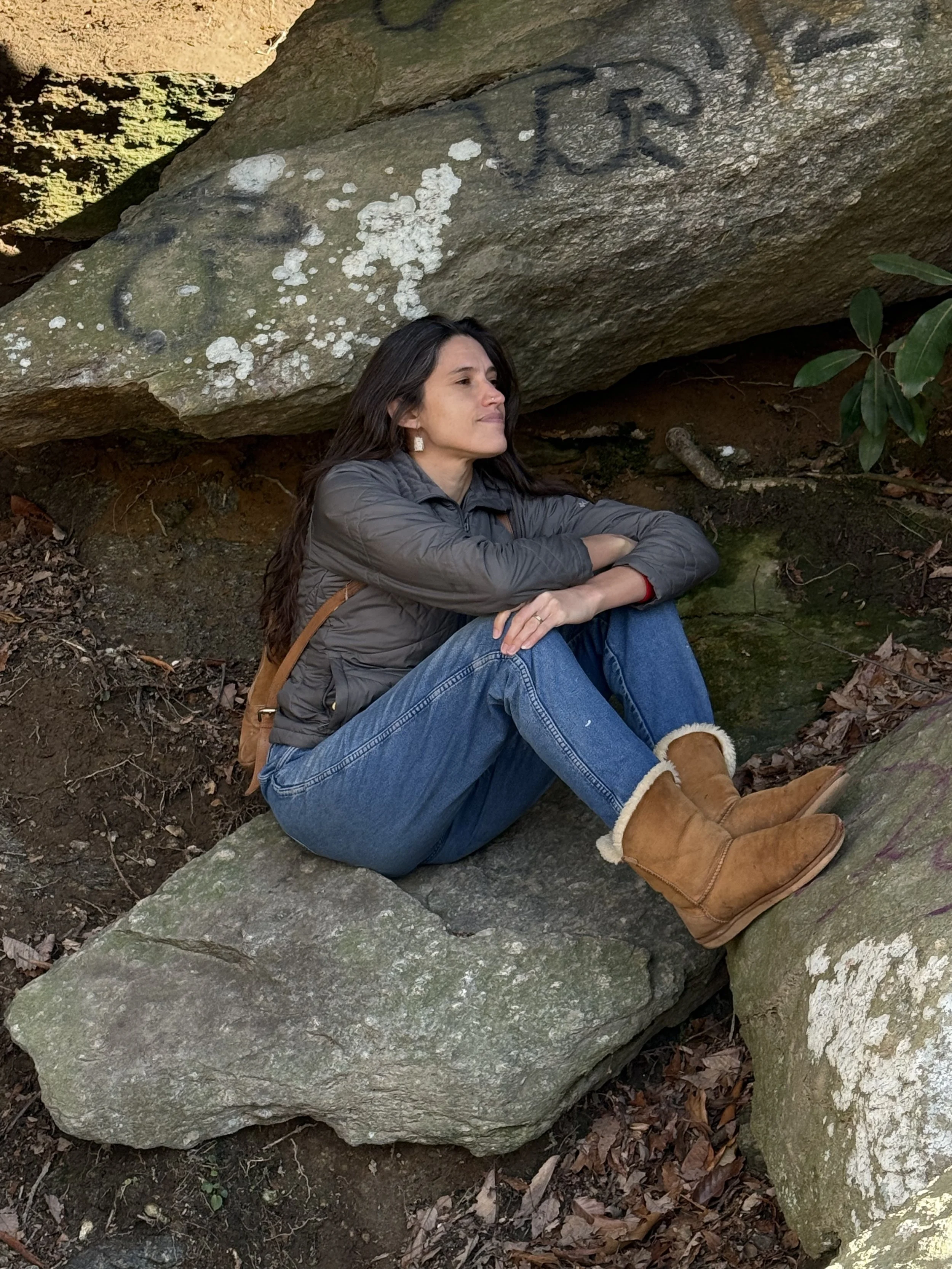A woman with long dark hair sitting on a large rock in a wooded area, resting her arms on her knees and looking to the side, wearing a gray jacket, blue jeans, and tan boots.