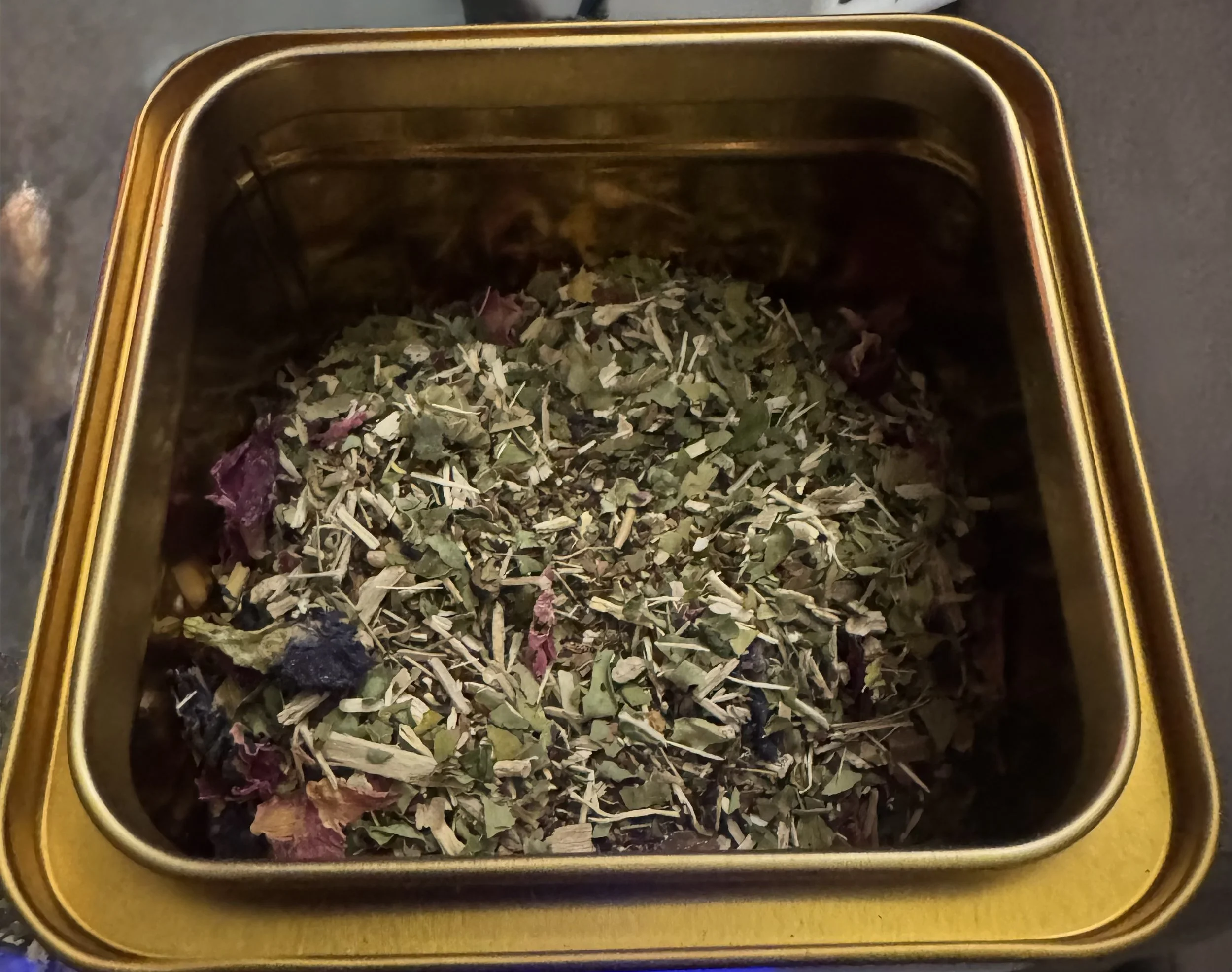 Golden tin container filled with dried herbal tea leaves and flowers.