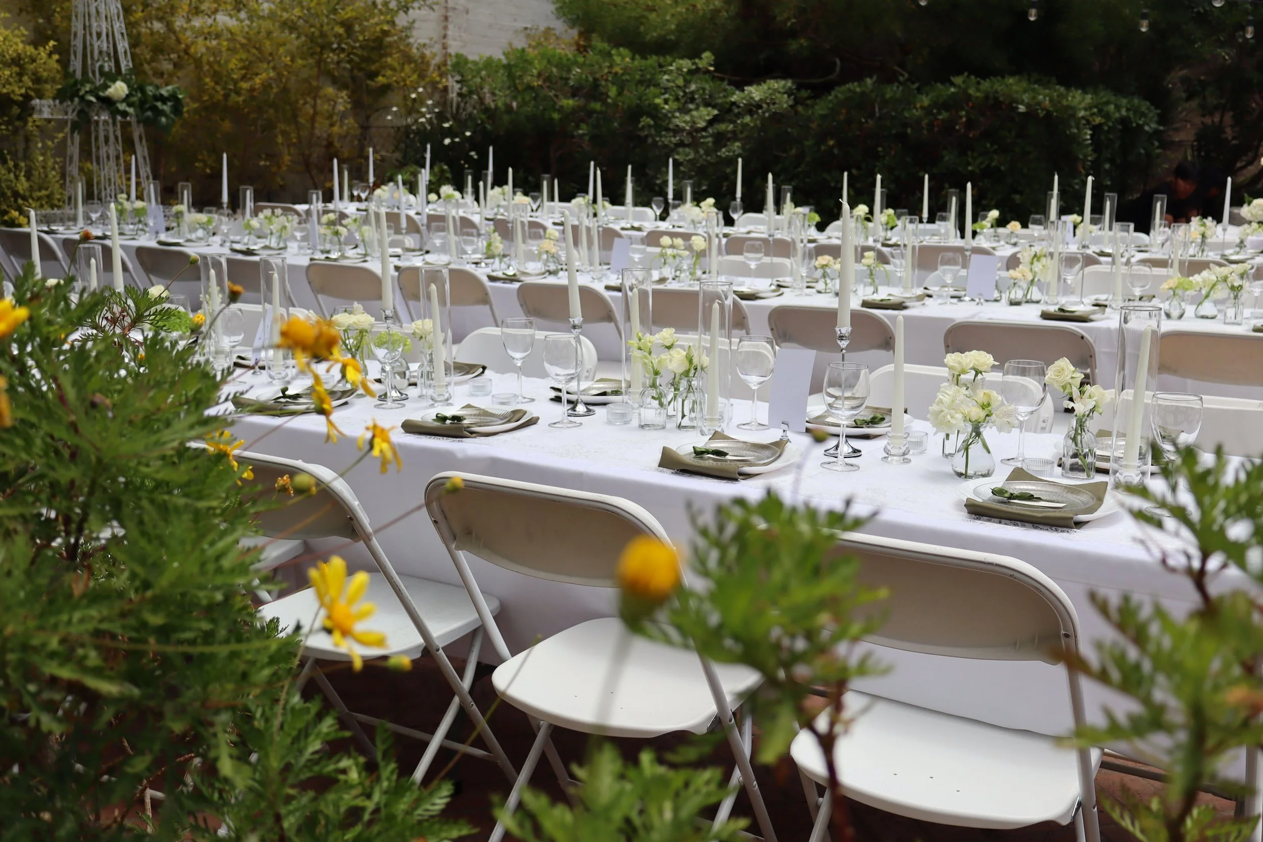 Long outdoor banquet table set for an event, with white tablecloths, floral centerpieces, plates, glasses, and taper candles, surrounded by white folding chairs, and surrounded by greenery.