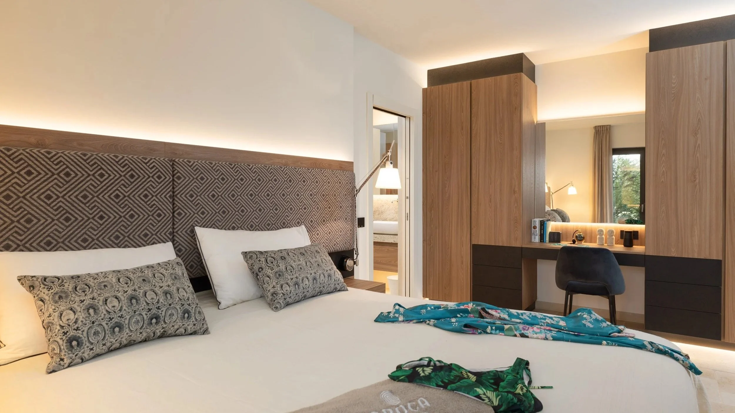 A modern hotel room with a bed, patterned pillows, a headboard, a desk with books and decor, beige walls, and a window with curtains.