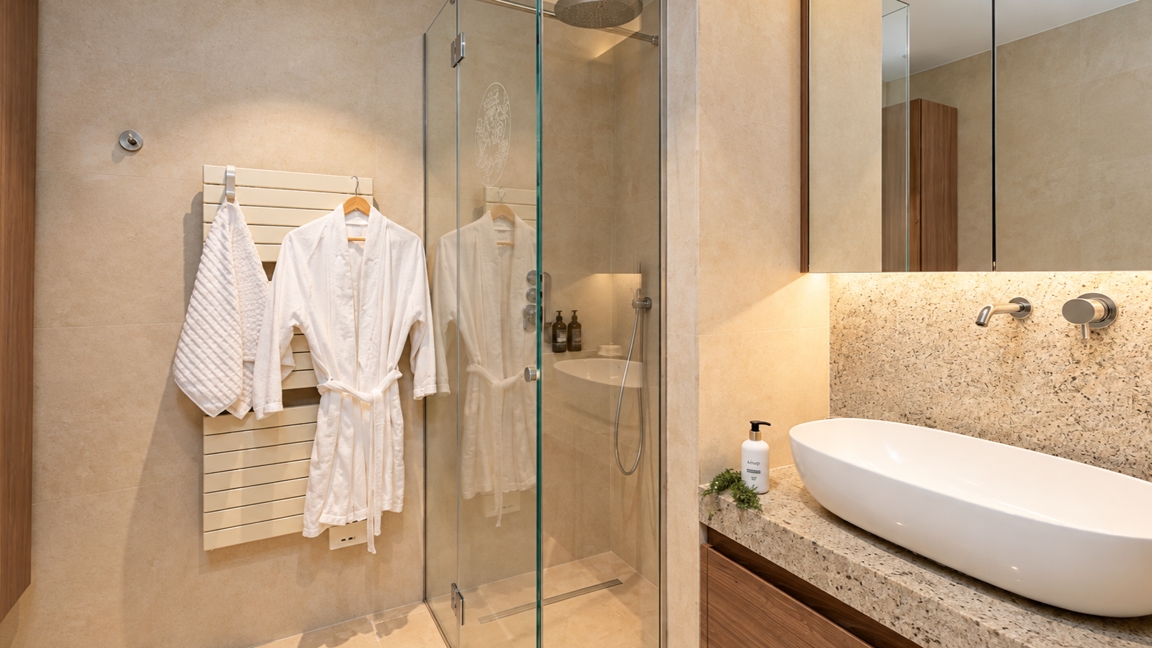 Modern bathroom with a glass shower, white bathrobe and towel hanging on a wall radiator, a large mirror over a granite countertop with a vessel sink, soap dispenser, and toiletries.