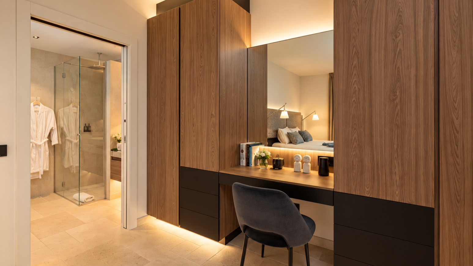 Hotel room with a dressing table, mirror, and chair, visible bathroom with glass shower enclosure, robes hanging inside, and towels on the floor.